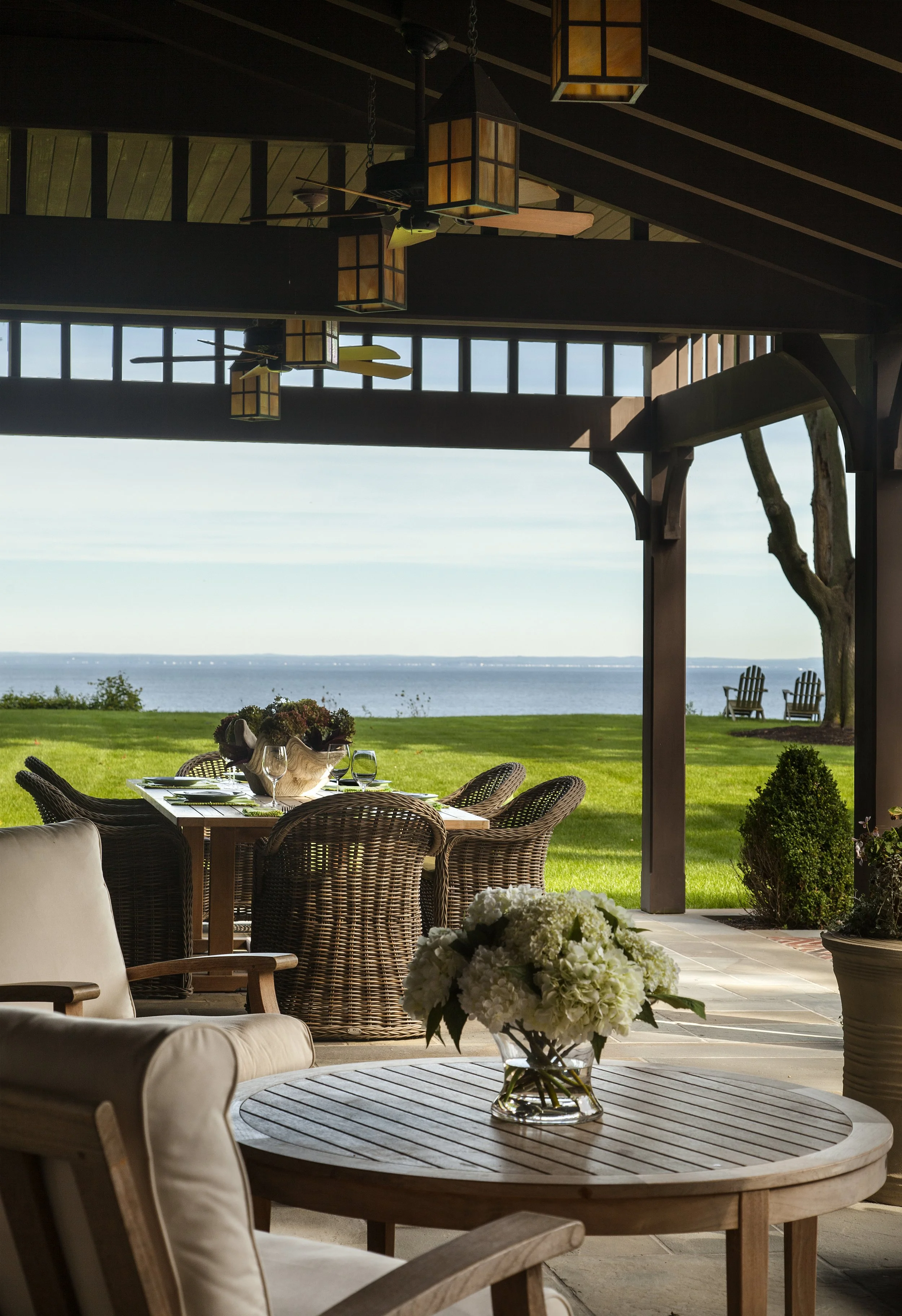An outdoor dining area with a table set for a meal, surrounded by wicker chairs, overlooking a lush green lawn and an ocean view. The scene includes a vase with white hydrangeas in the foreground, and two Adirondack chairs in the distance facing the 