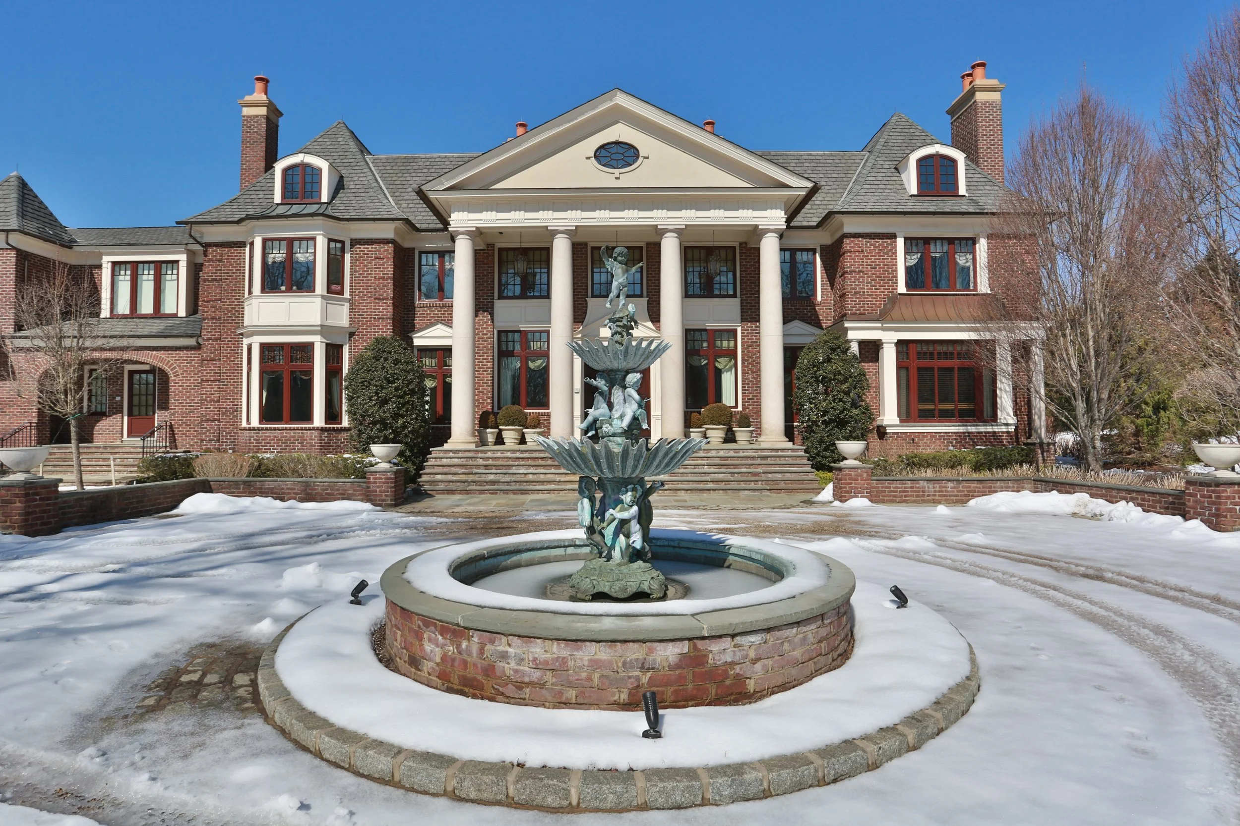 Large brick mansion with a fountain in front, snow on the ground, and a clear blue sky.