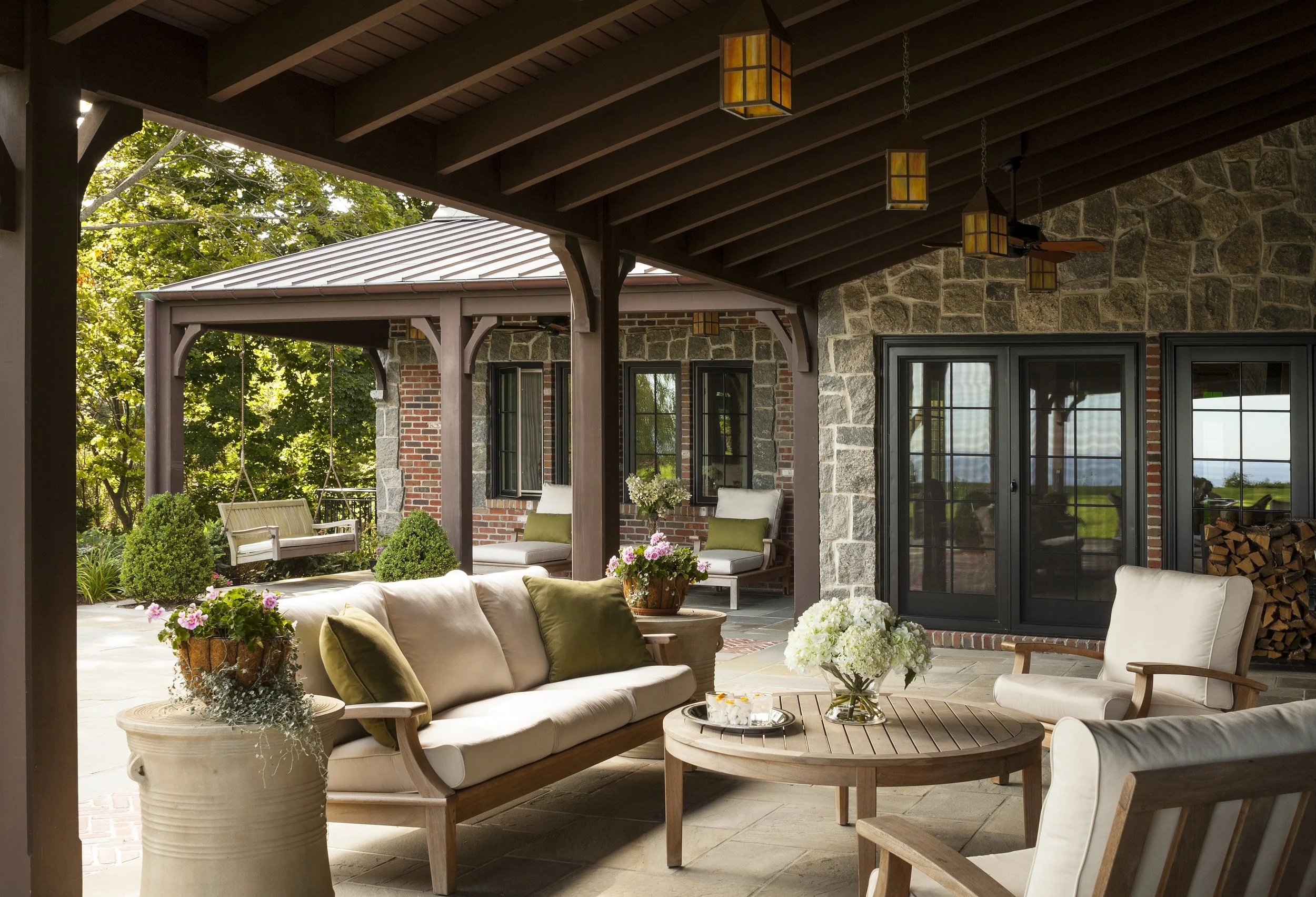Outdoor patio with beige cushioned furniture, potted flowers, a round wooden table, decorative lantern ceiling lights, and a brick and stone house exterior with large windows.