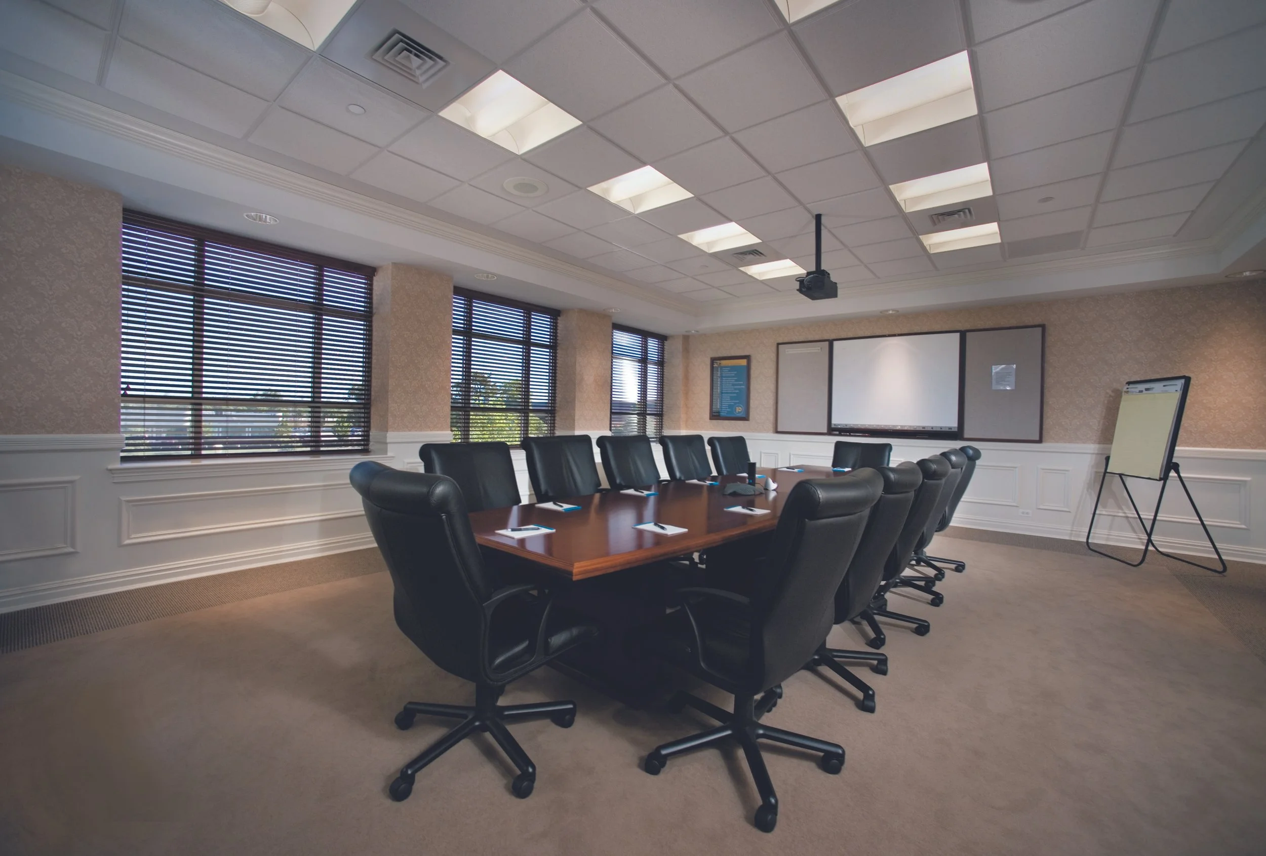 Empty conference room with a long wooden table, ten black leather office chairs, windows with blinds, a whiteboard, and a ceiling-mounted projector.