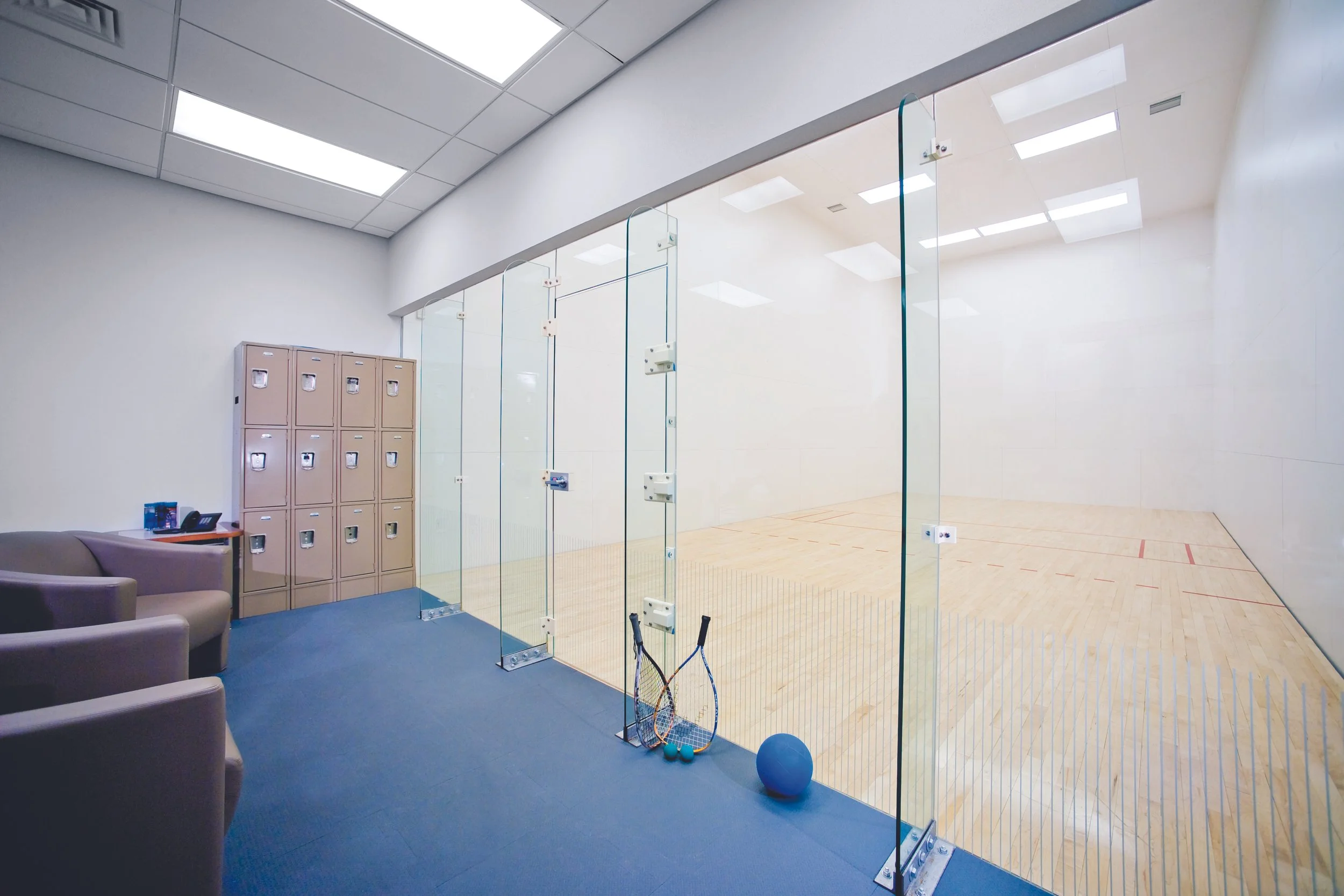 Indoor squash court with a glass back wall, lockers in corner, and sports equipment including tennis rackets, balls, and a blue exercise ball in front of the court.