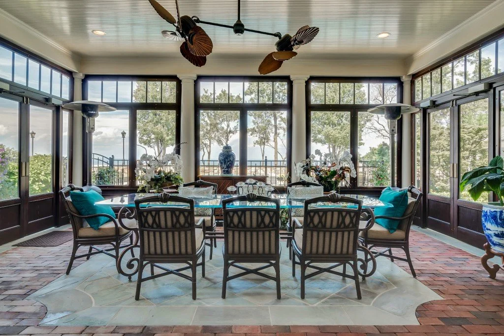 A bright enclosed sunroom with large windows, a glass-top dining table surrounded by eight chairs with striped cushions and turquoise pillows, decorated with floral centerpieces and vases, with outdoor scenery visible through the windows and a ceilin