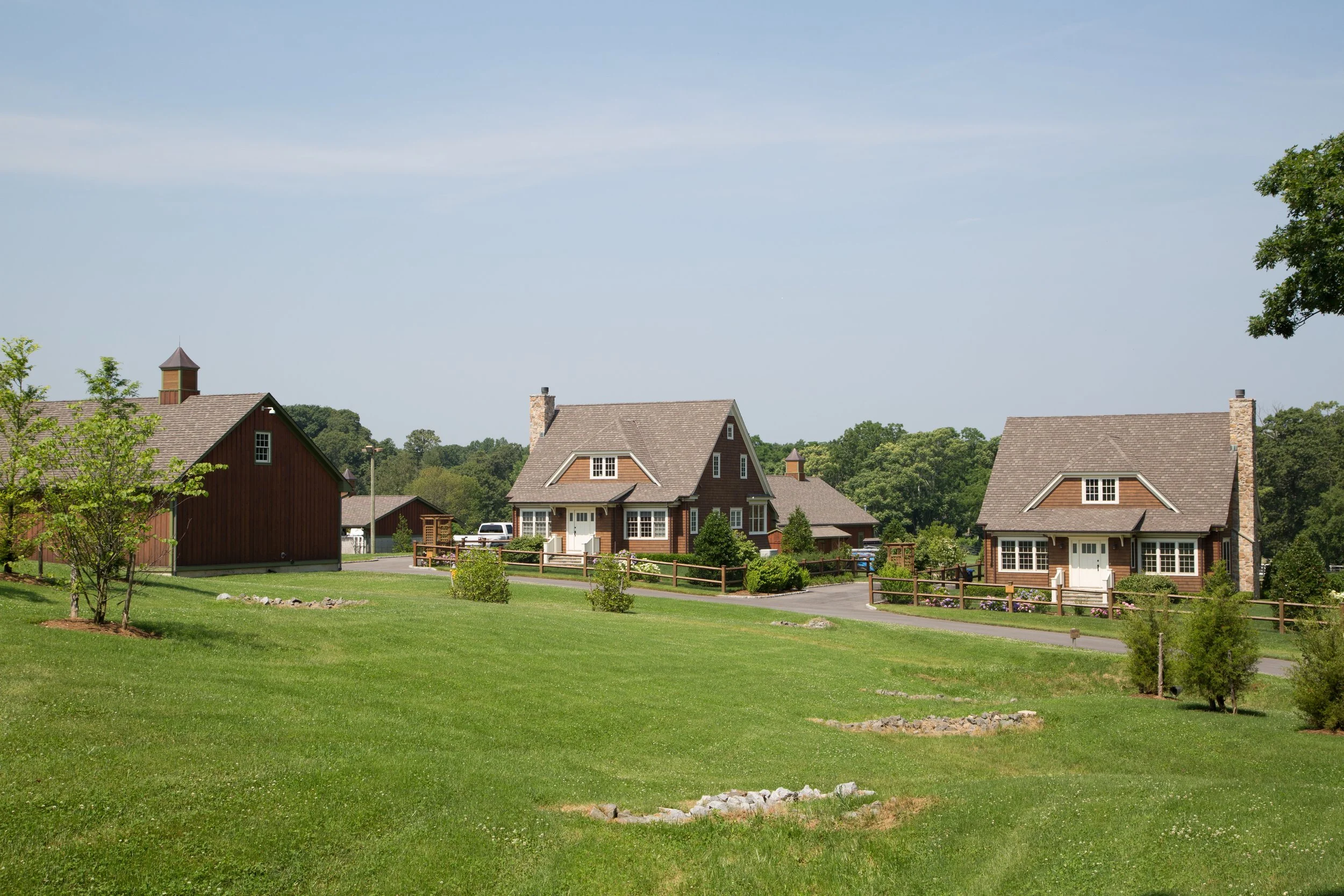 A peaceful suburban neighborhood with two large houses, well-maintained lawns, trees, and a wooden fence, under a clear blue sky.