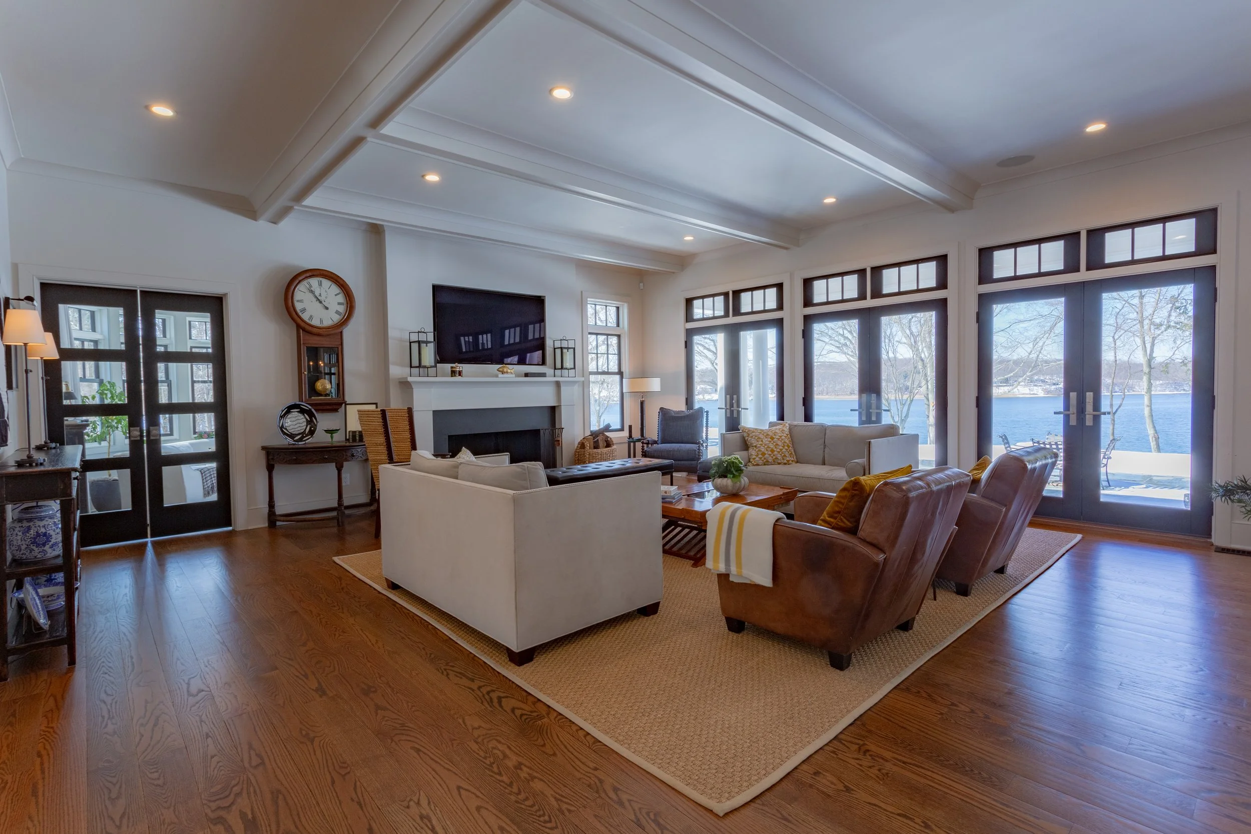 Bright living room with hardwood floors, white walls, and large windows overlooking a lake. Includes sofas, armchairs, a fireplace, and a wall-mounted TV.