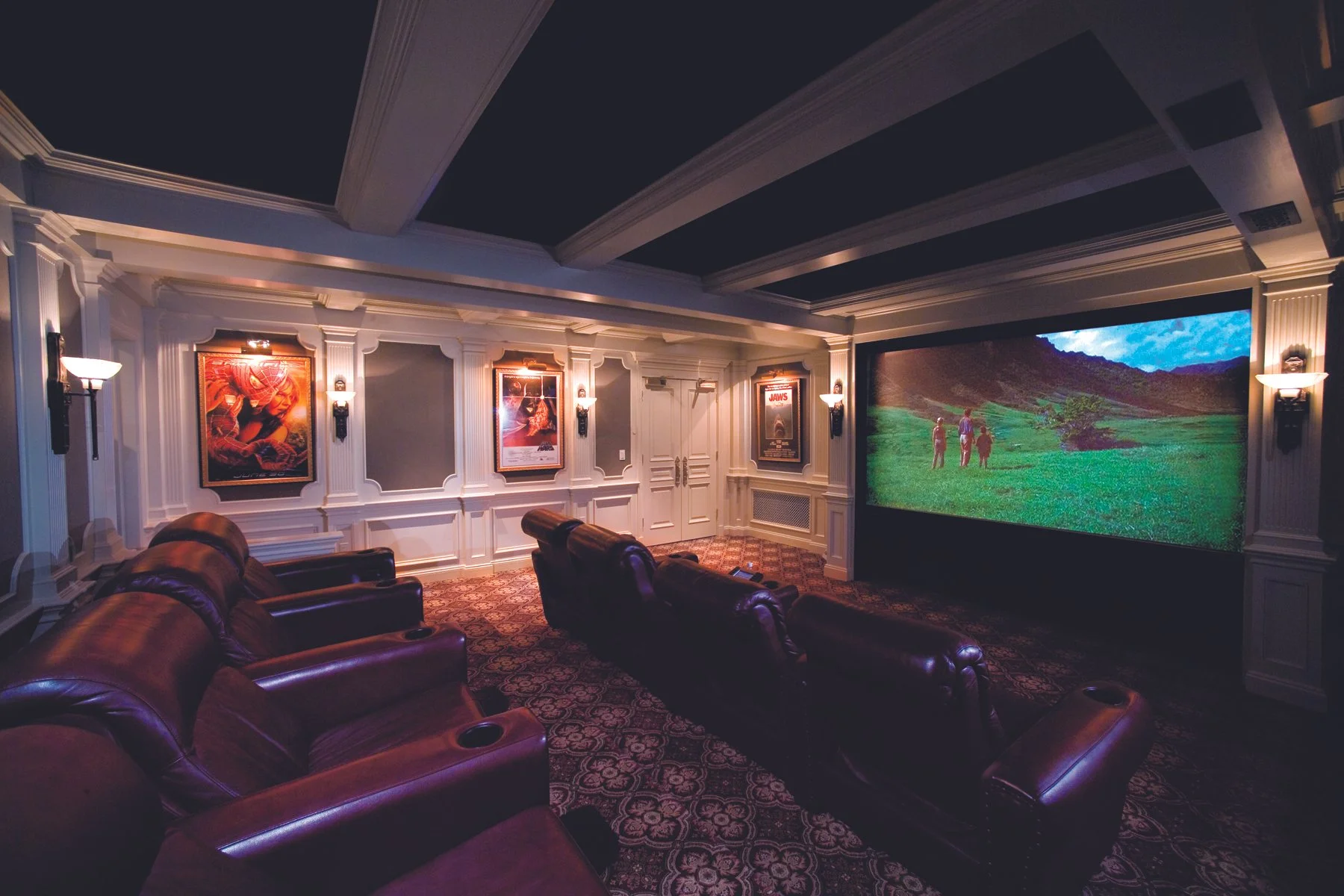A luxurious home theater with four leather recliner chairs facing a large screen displaying a scenic green landscape with three people walking. The walls are decorated with framed movie posters and ornate white paneling, with wall sconces providing a