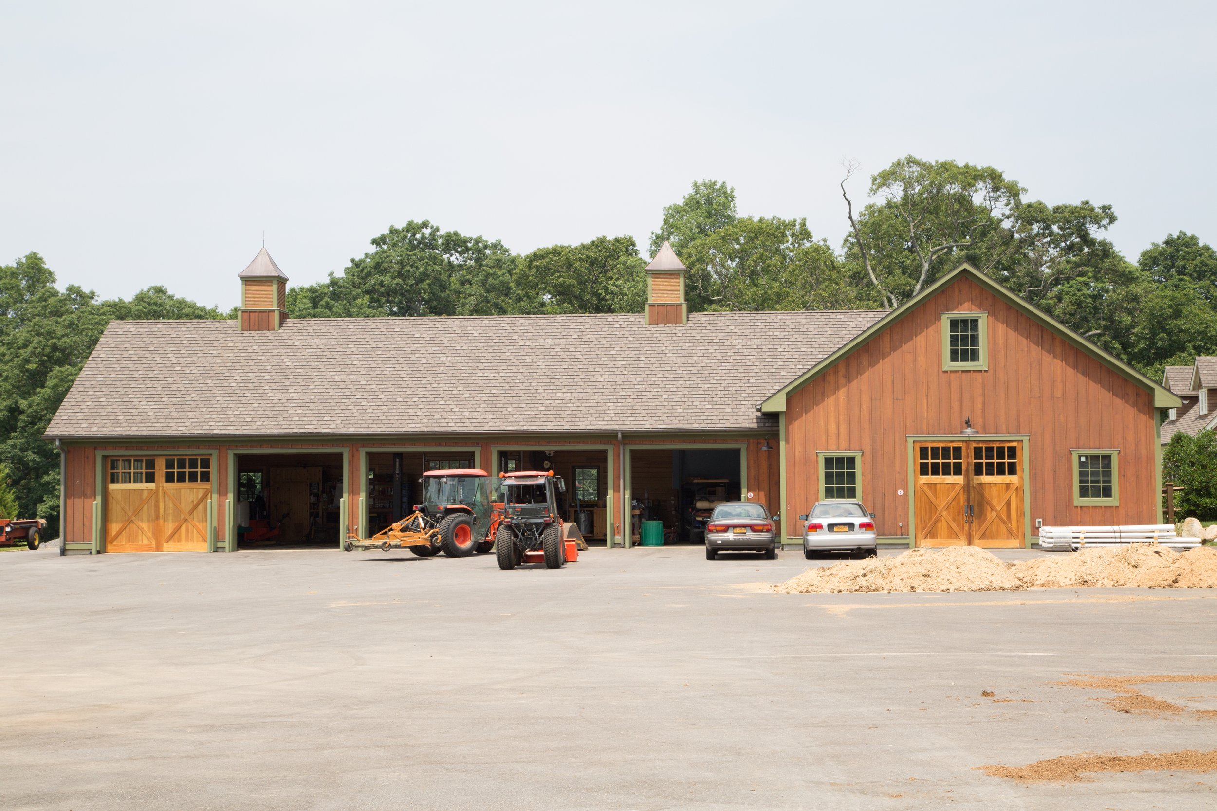 A large barn with green-framed windows and doors, a shingled roof, and two small towers, situated in a rural area with trees in the background and cars parked outside.