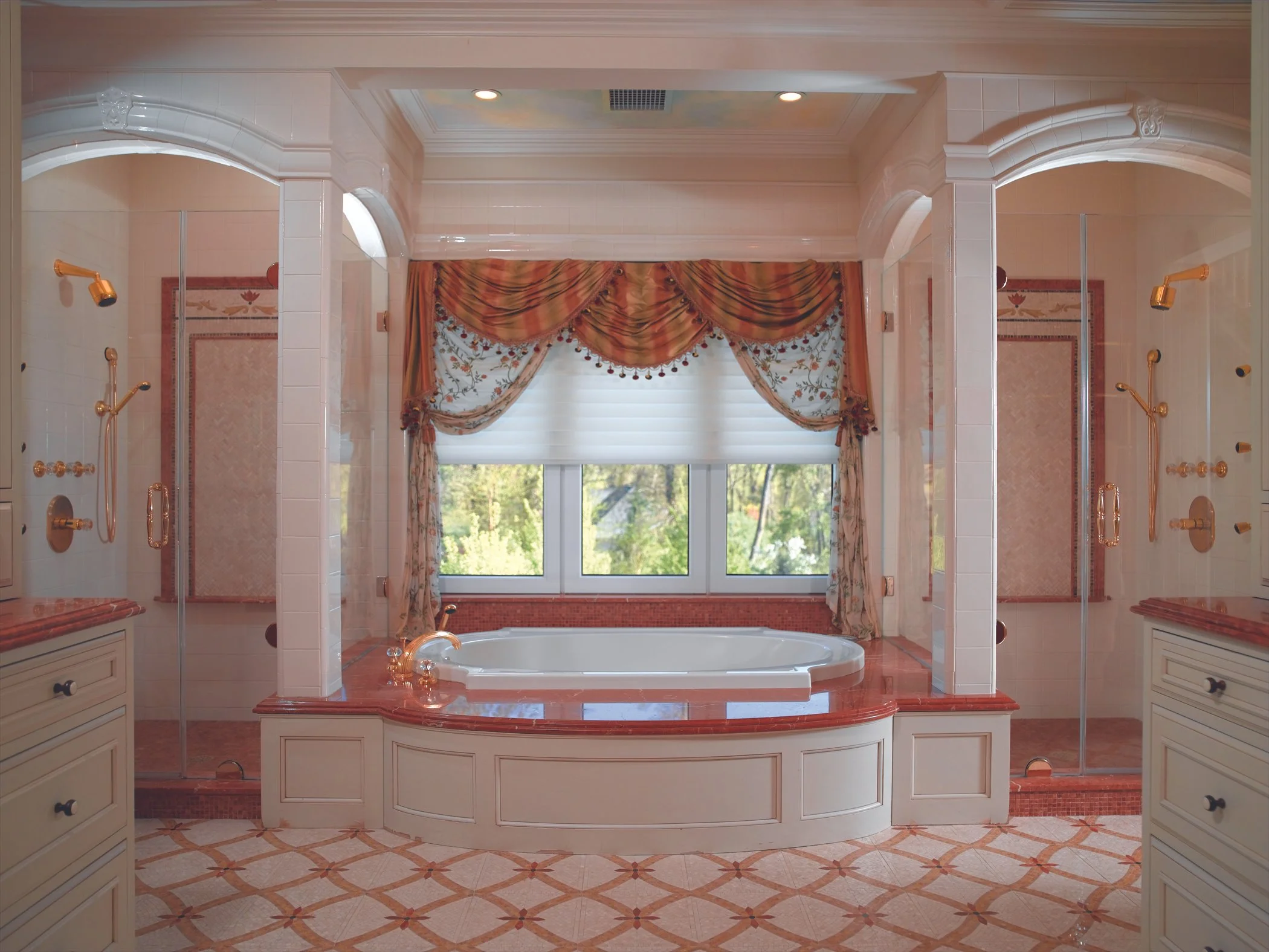 A spacious bathroom with a bathtub in front of a large window with floral curtains, surrounded by divided showers and cream-colored cabinetry.