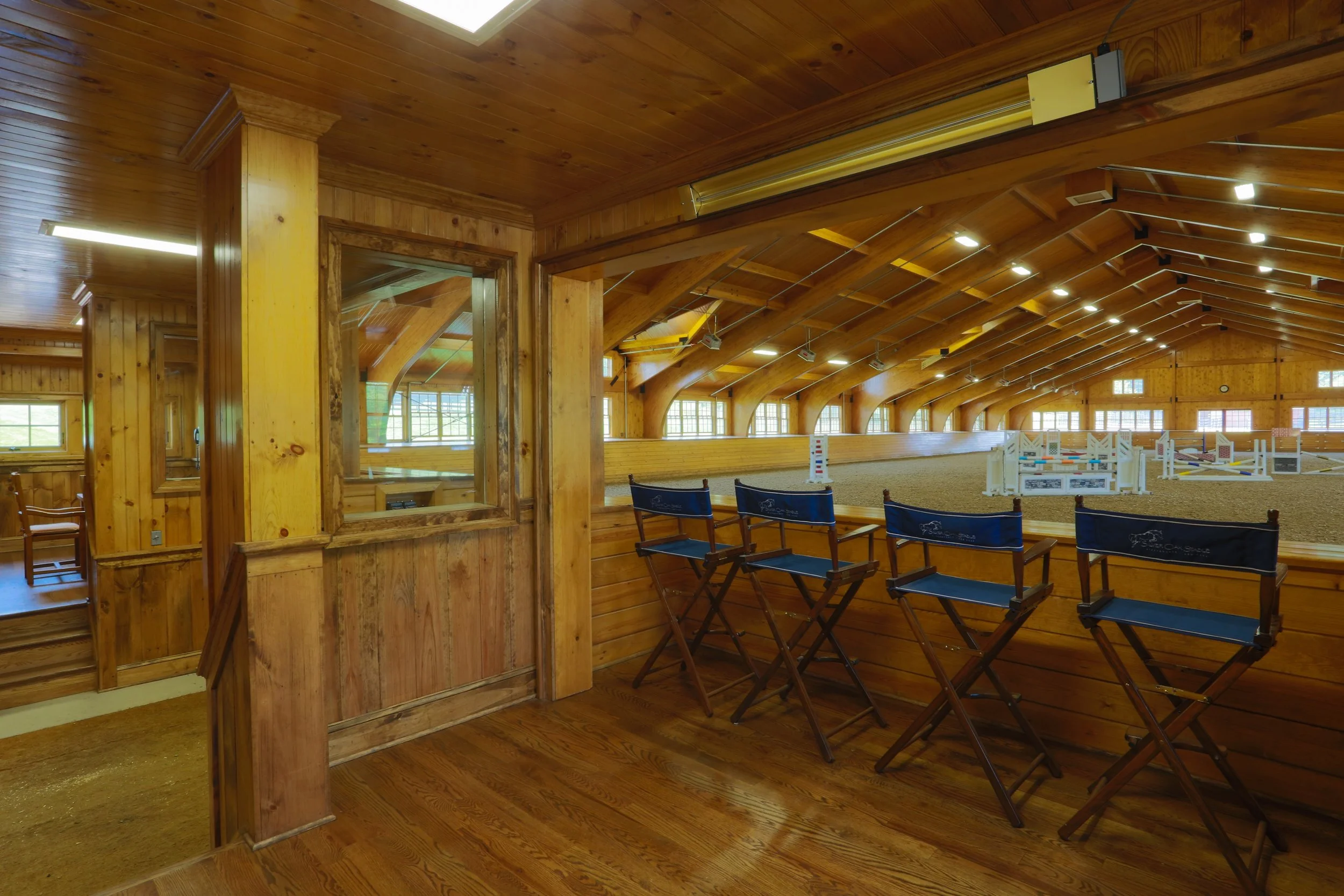 View of an indoor horse riding arena with a wooden interior, showing jump obstacles and seating chairs.