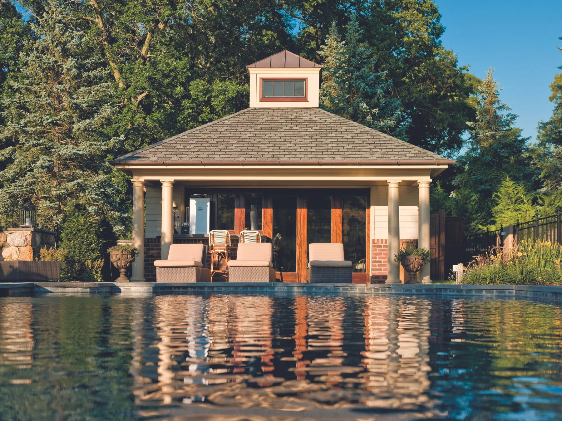 Backyard with a swimming pool, patio furniture, and a small building with sliding glass doors and a cupola, surrounded by trees.