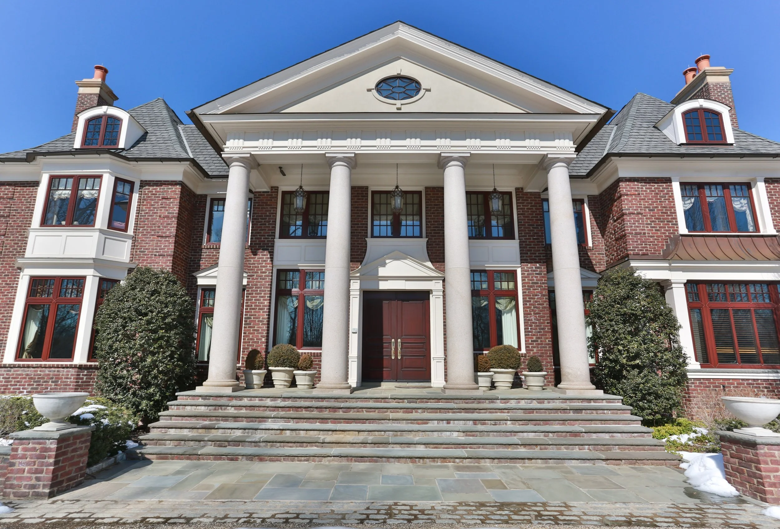 Front view of a large brick house with four tall white columns, a staircase, and red-framed windows leading up to a brown double door under a gable roof.