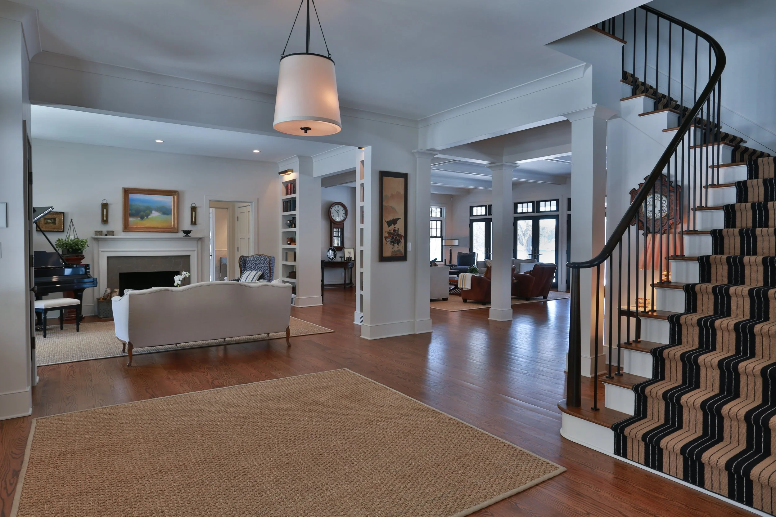 View of a spacious, well-lit living room with hardwood floors, white walls, a staircase with striped carpet, and multiple seating areas with chairs and sofas.