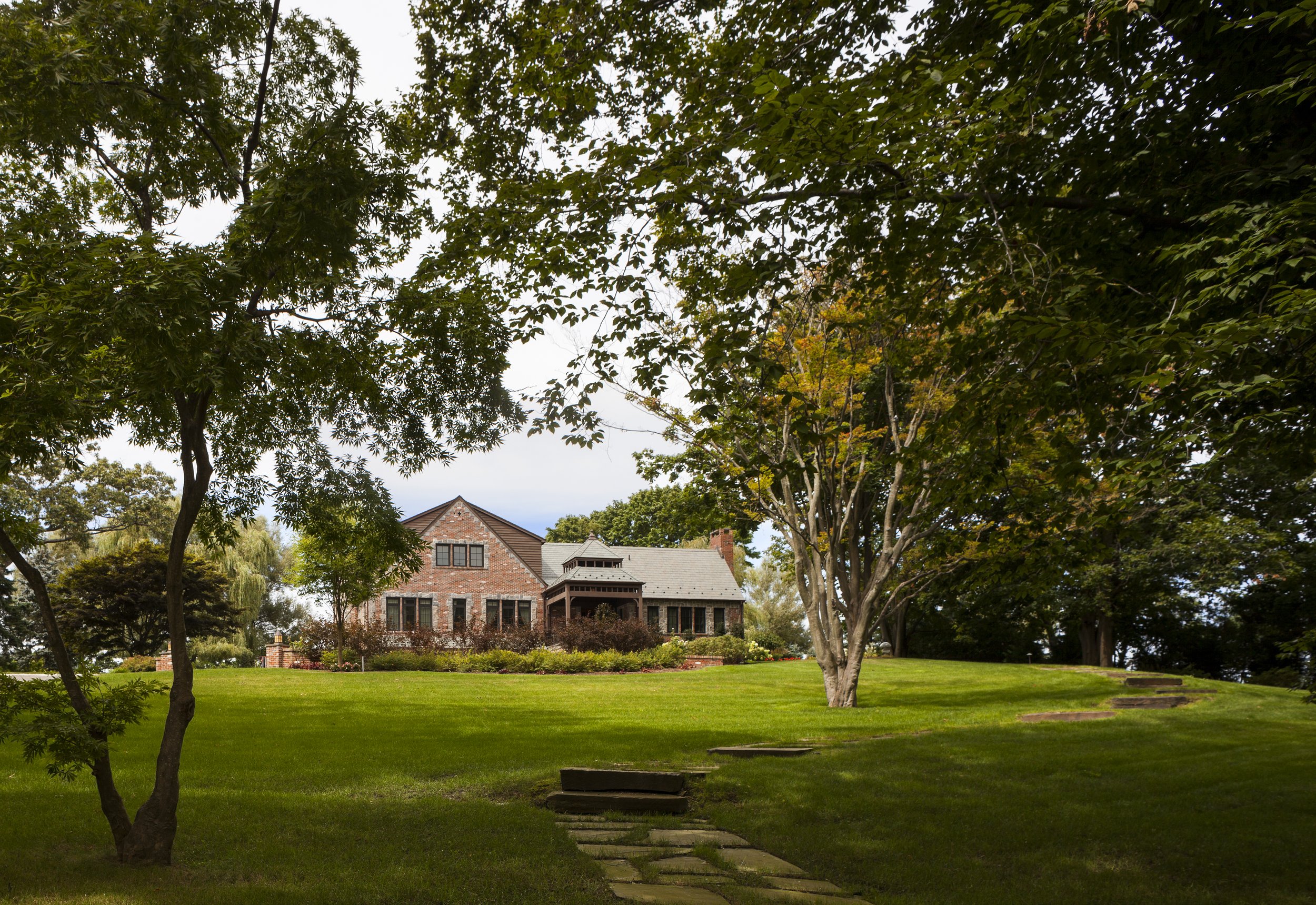 A large brick house on a hillview with a well-maintained lawn and trees surrounding it.