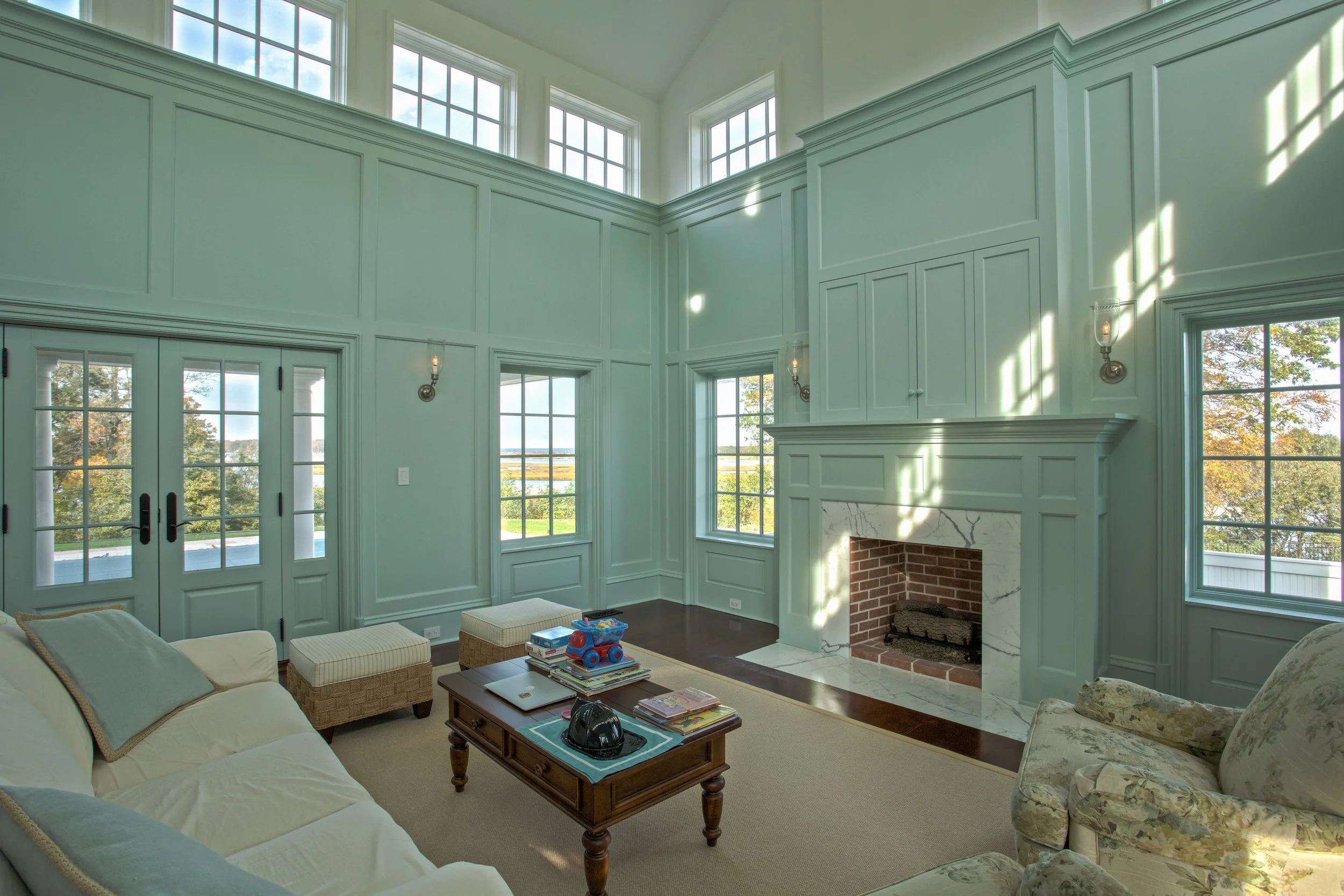 Bright living room with pale green walls, multiple windows, a brick and marble fireplace, beige and floral upholstered furniture, wooden coffee table, and sunlight streaming through windows.