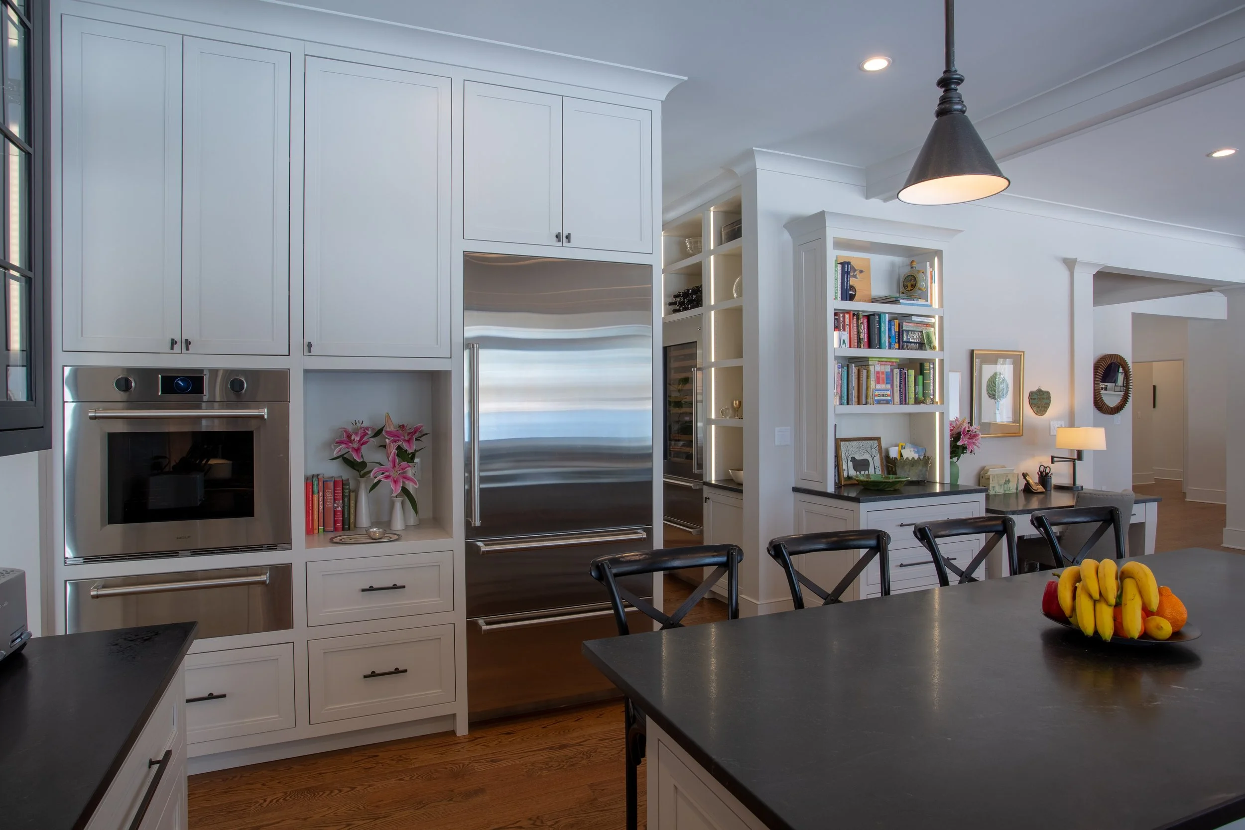 Kitchen with white cabinets, stainless steel refrigerator, built-in oven, open shelves, black countertop island, a bowl of bananas and fruit, and a desk area with books and decorative items.