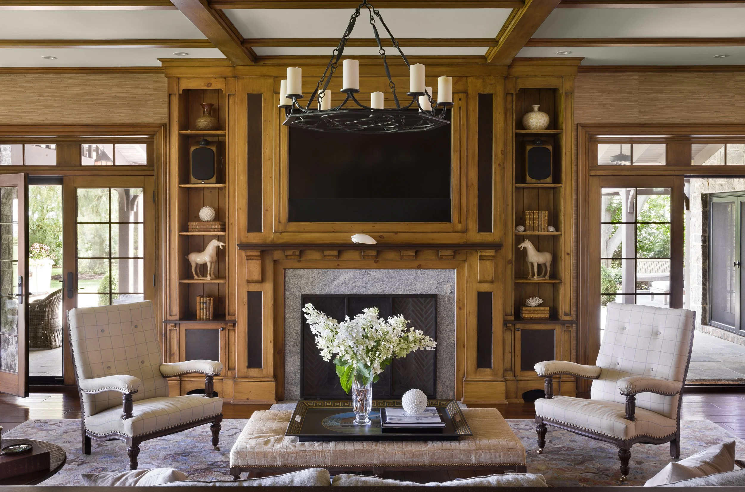 Living room with wooden wall paneling, a fireplace with a black screen, two beige armchairs, a coffee table with a vase of flowers, and large windows showing an outdoor patio.