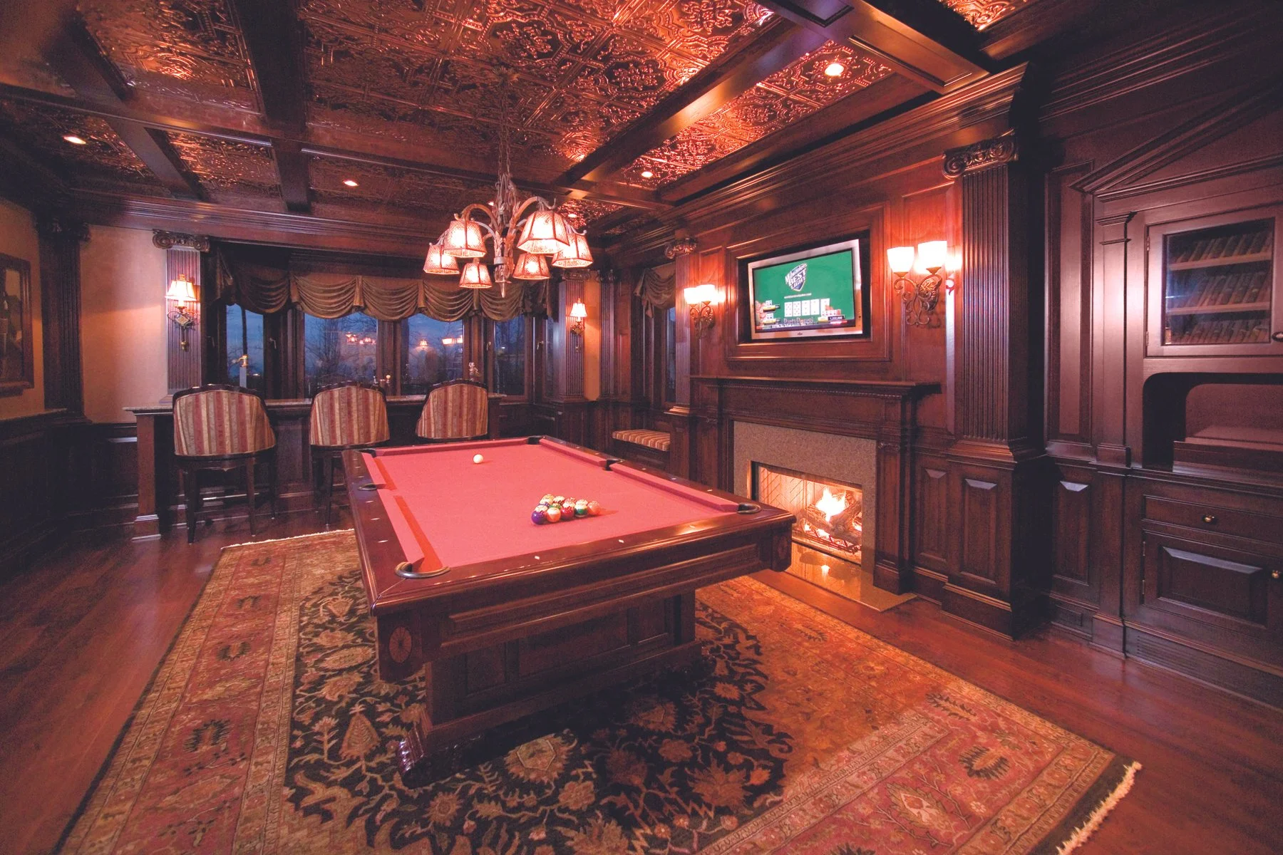 A luxurious wood-paneled game room with a pool table, a fireplace with a fire, a flat-screen TV showing a card game, and a chandelier with multiple lamps, decorated with ornate ceiling details and traditional rugs.