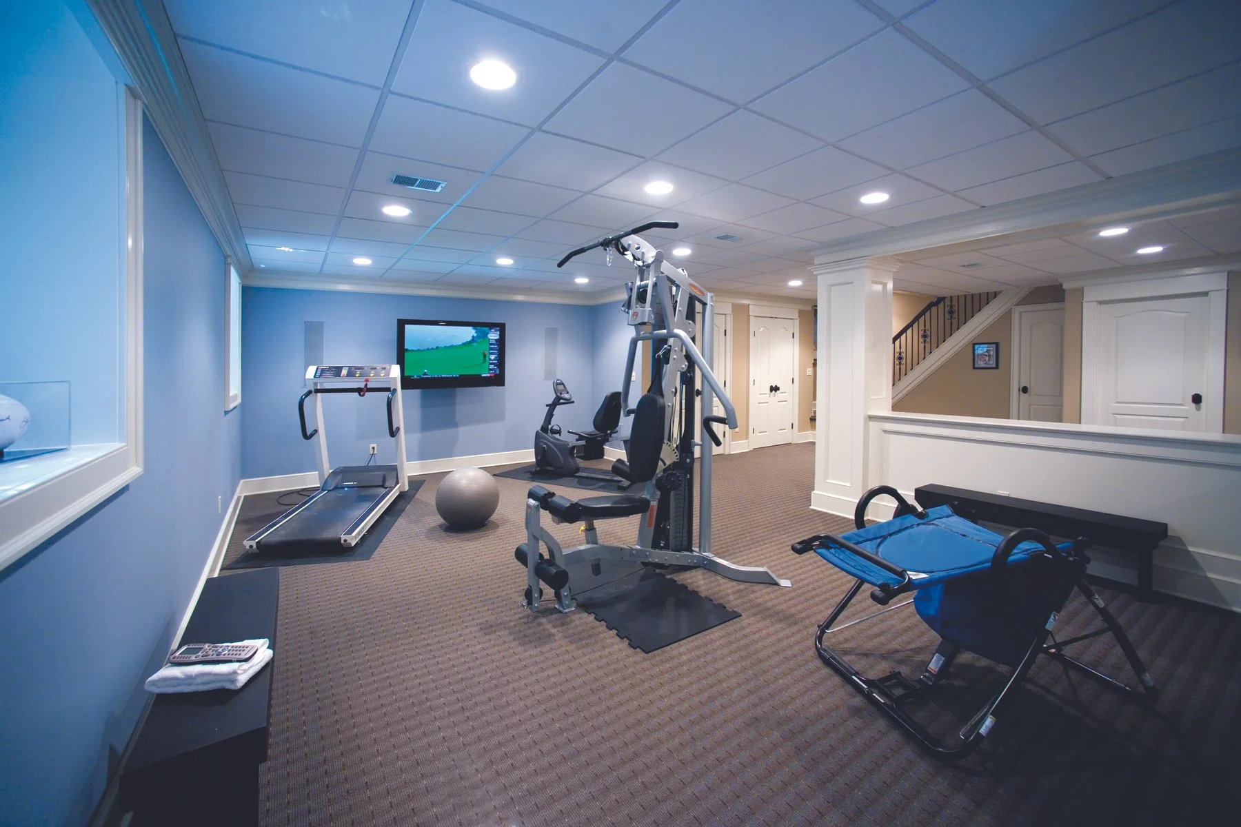 Home gym with treadmill, exercise bike, multi-station gym equipment, stability ball, sled, and a wall-mounted TV in a basement, with white walls and a brown carpeted floor.