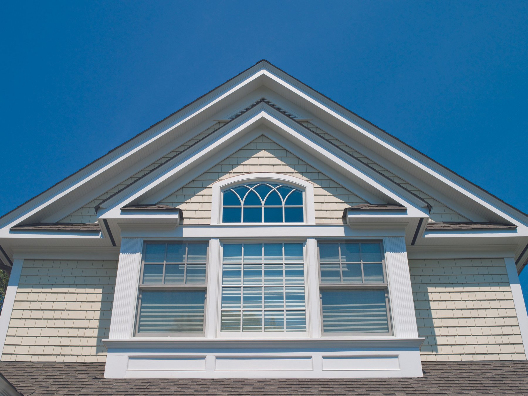 Close-up of the upper part of a house with beige siding, white trim, large window with shutters, and a steep gabled roof under a clear blue sky.