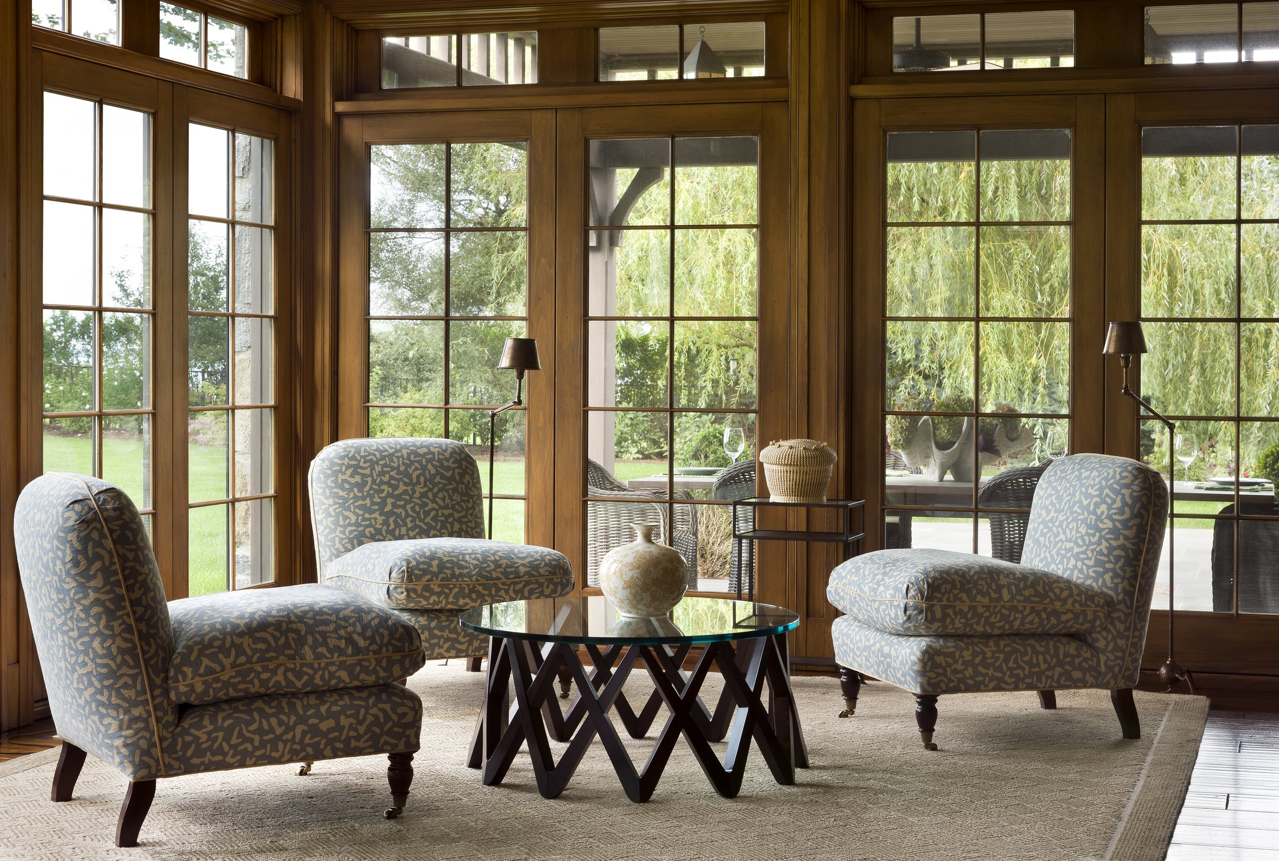 Living room with large wooden-framed windows overlooking a green garden, featuring four patterned armchairs around a glass-top coffee table, with decorative objects and a woven basket on a side table.