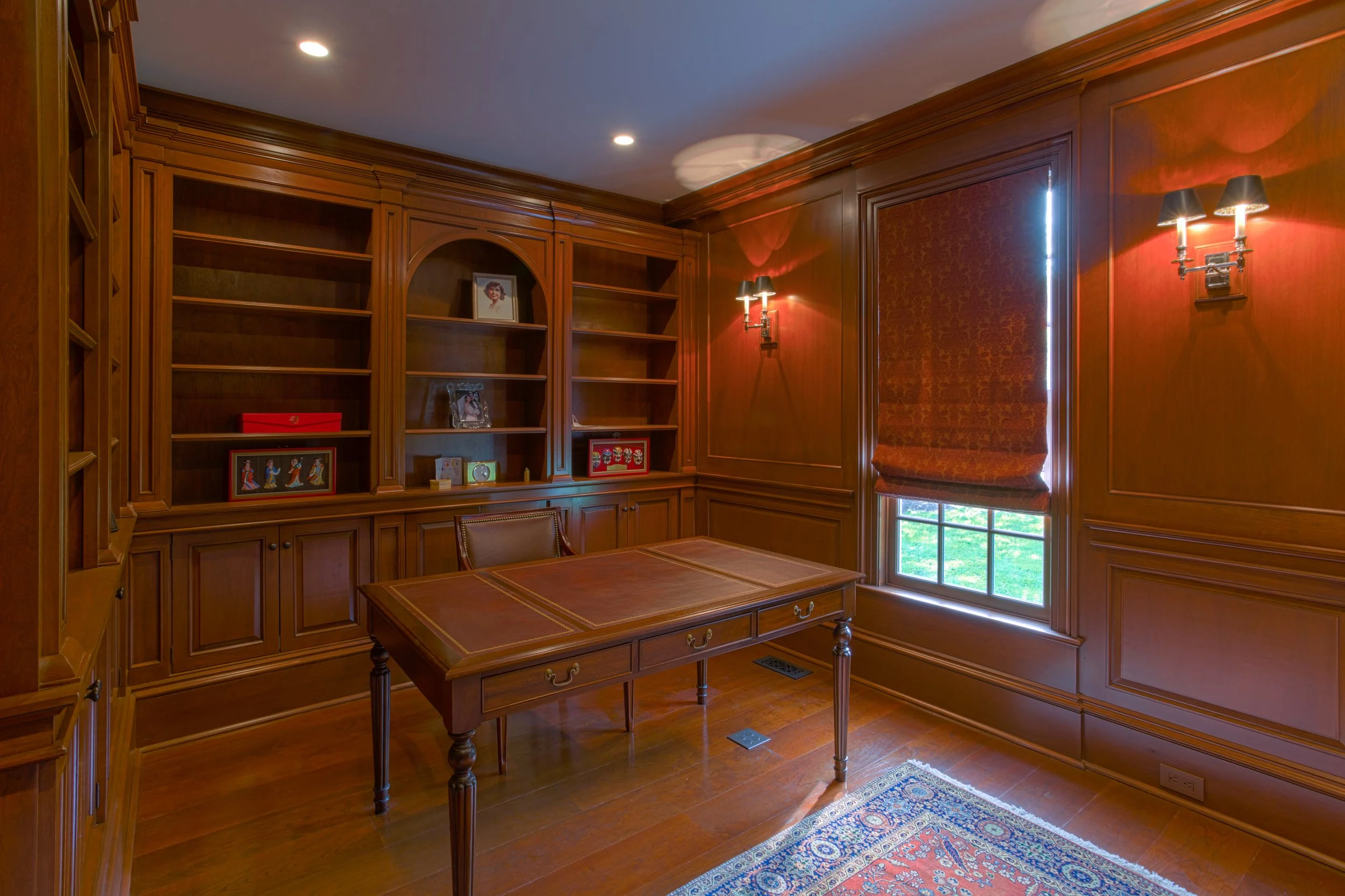 A room with wood-paneled walls, a wooden desk, a leather chair, built-in wooden bookshelves, a window with a red patterned shade, and wall-mounted lamps.