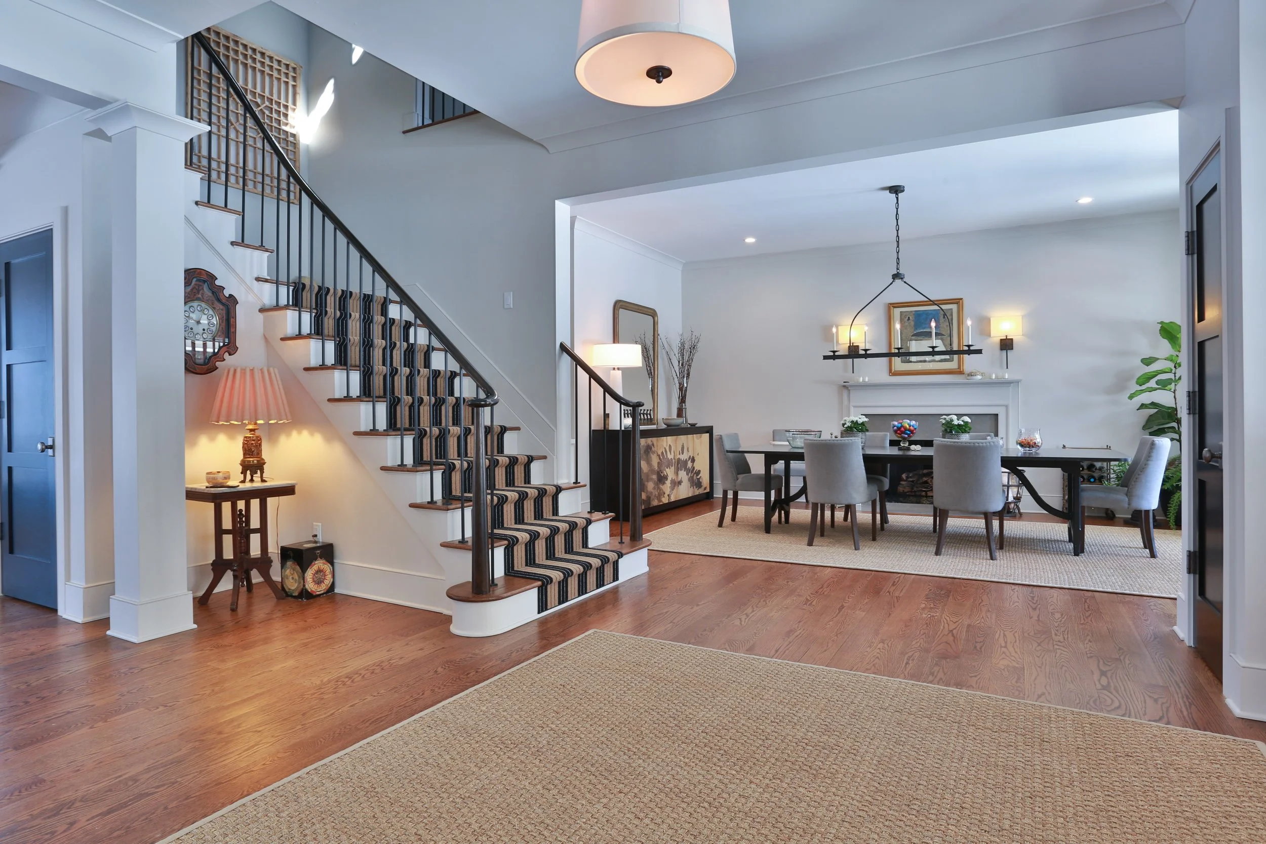 Interior of a home with a staircase on the left and a dining area on the right. The staircase has a striped carpet runner, black railing, and wooden steps. The dining area features a black table with eight gray chairs, a fireplace with framed art and