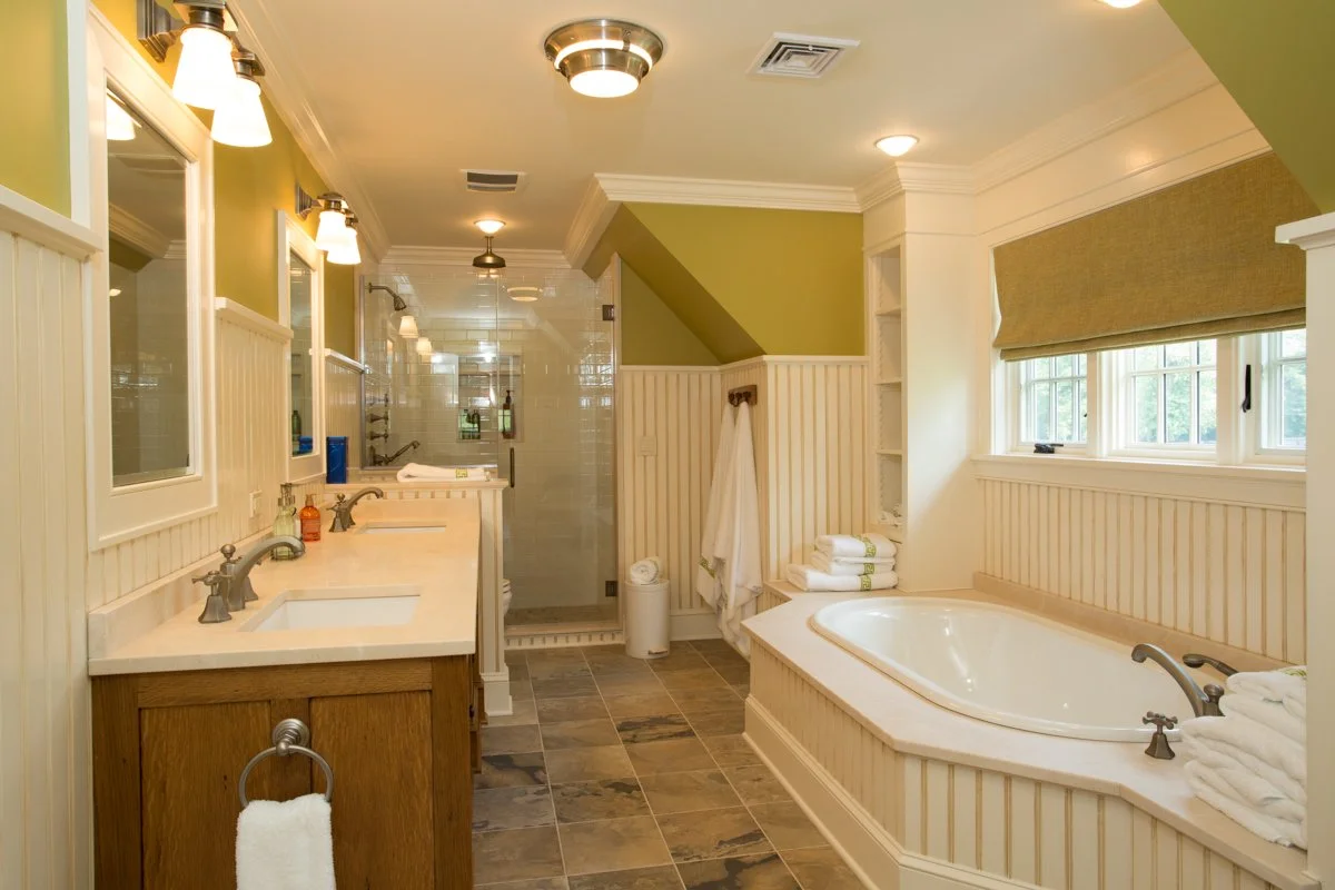 A spacious bathroom with dual sinks, a bathtub beneath a window, and a walk-in shower in the background.