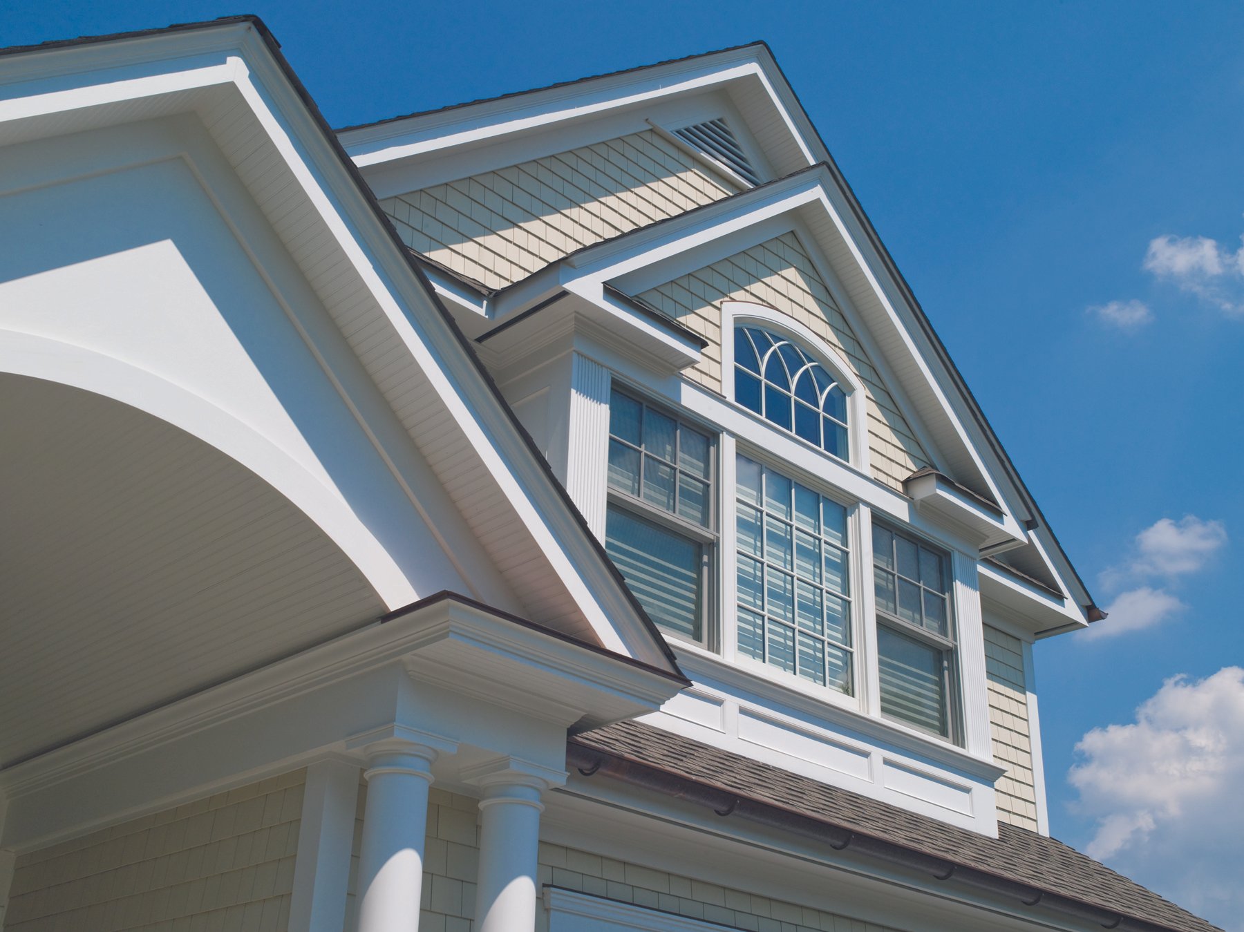 Close-up of a modern house's exterior showing decorative windows, columns, and pitched roof against a blue sky.