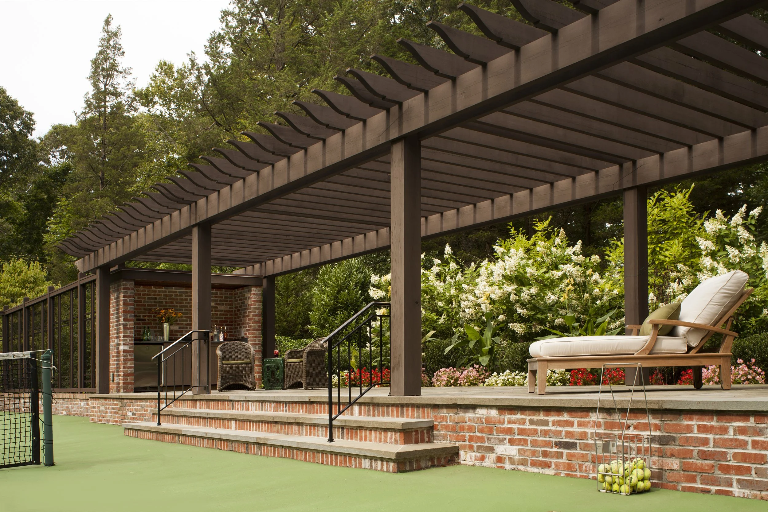 Patio area with a wooden pergola, brick wall, wicker chairs, a cushioned lounge chair, a side table, a basket of tennis balls, and lush green foliage with white flowers in the background.