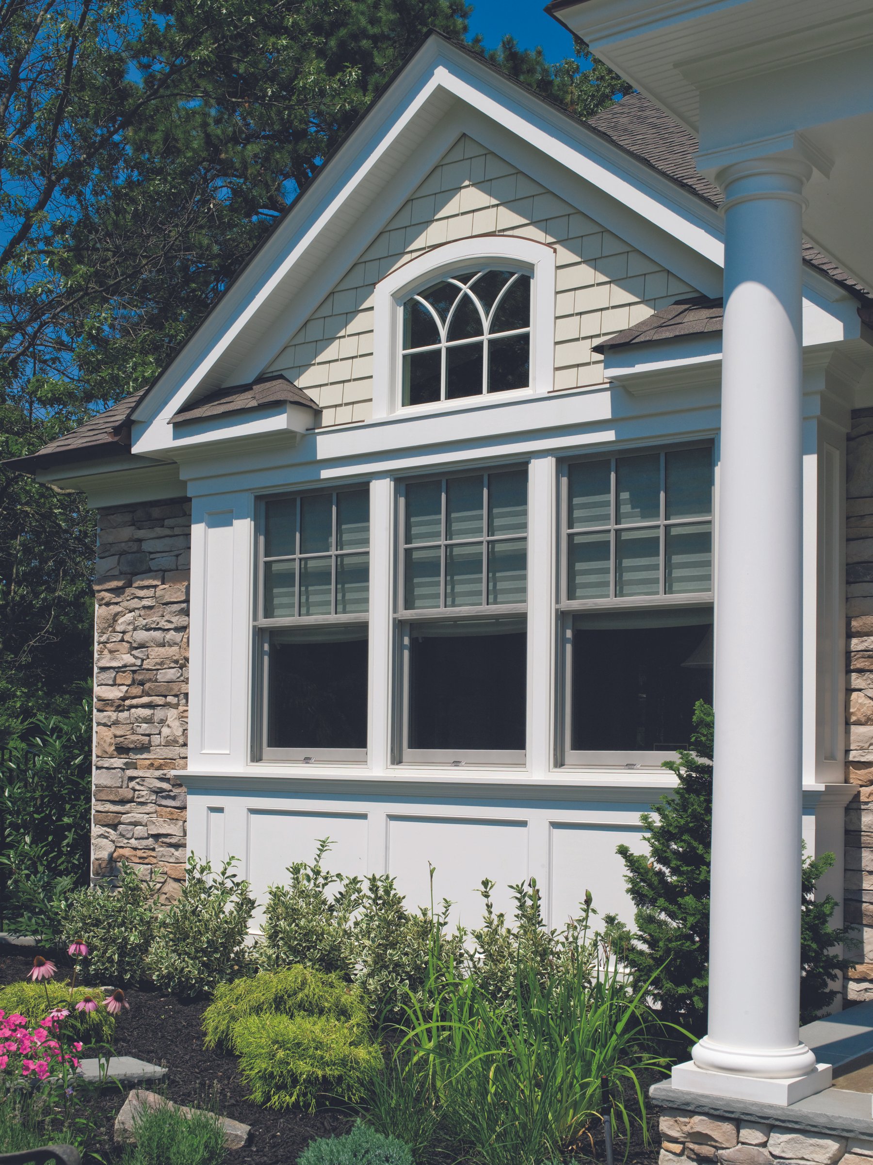 Front view of a house featuring a large window with multiple panes, a triangular dormer window with decorative arches, stone and siding exterior, and a white column on the porch, surrounded by a garden with various green plants and pink flowers.