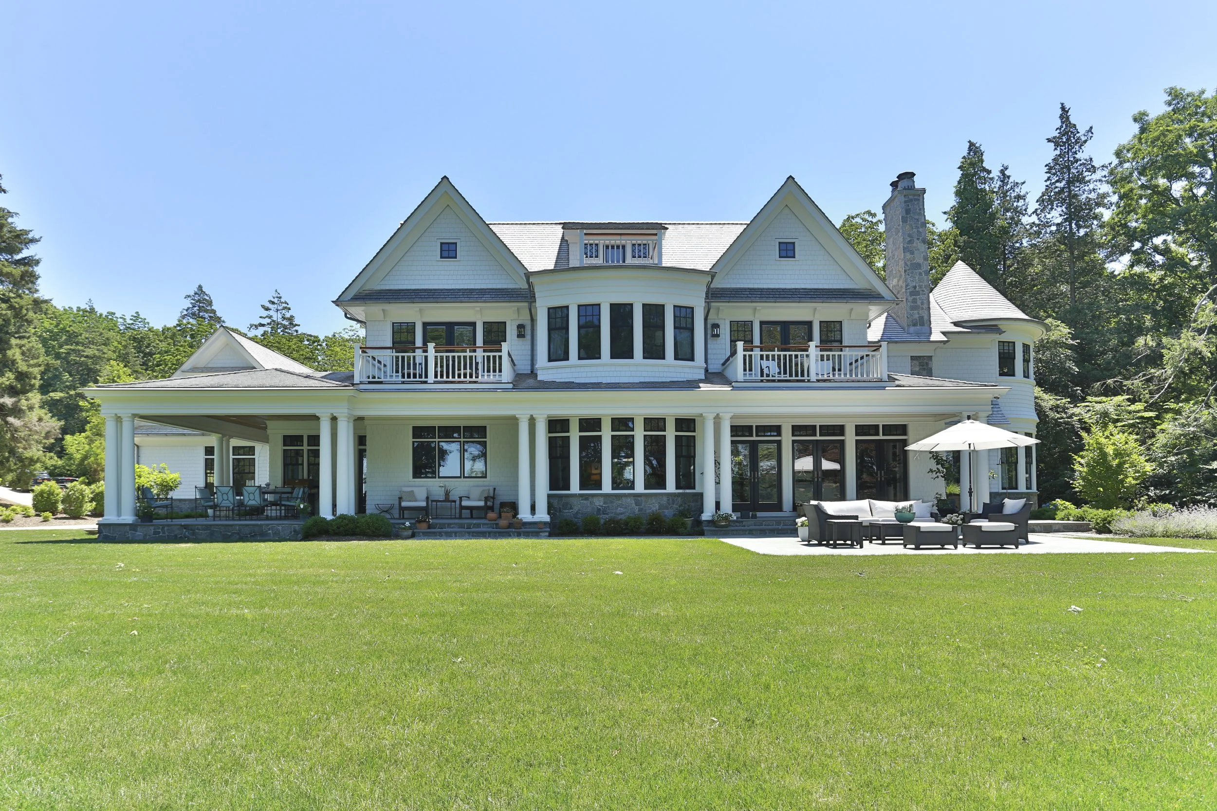 Large white multi-story house with wraparound porch, black window frames, and a chimney, surrounded by green lawn and trees under a clear blue sky.
