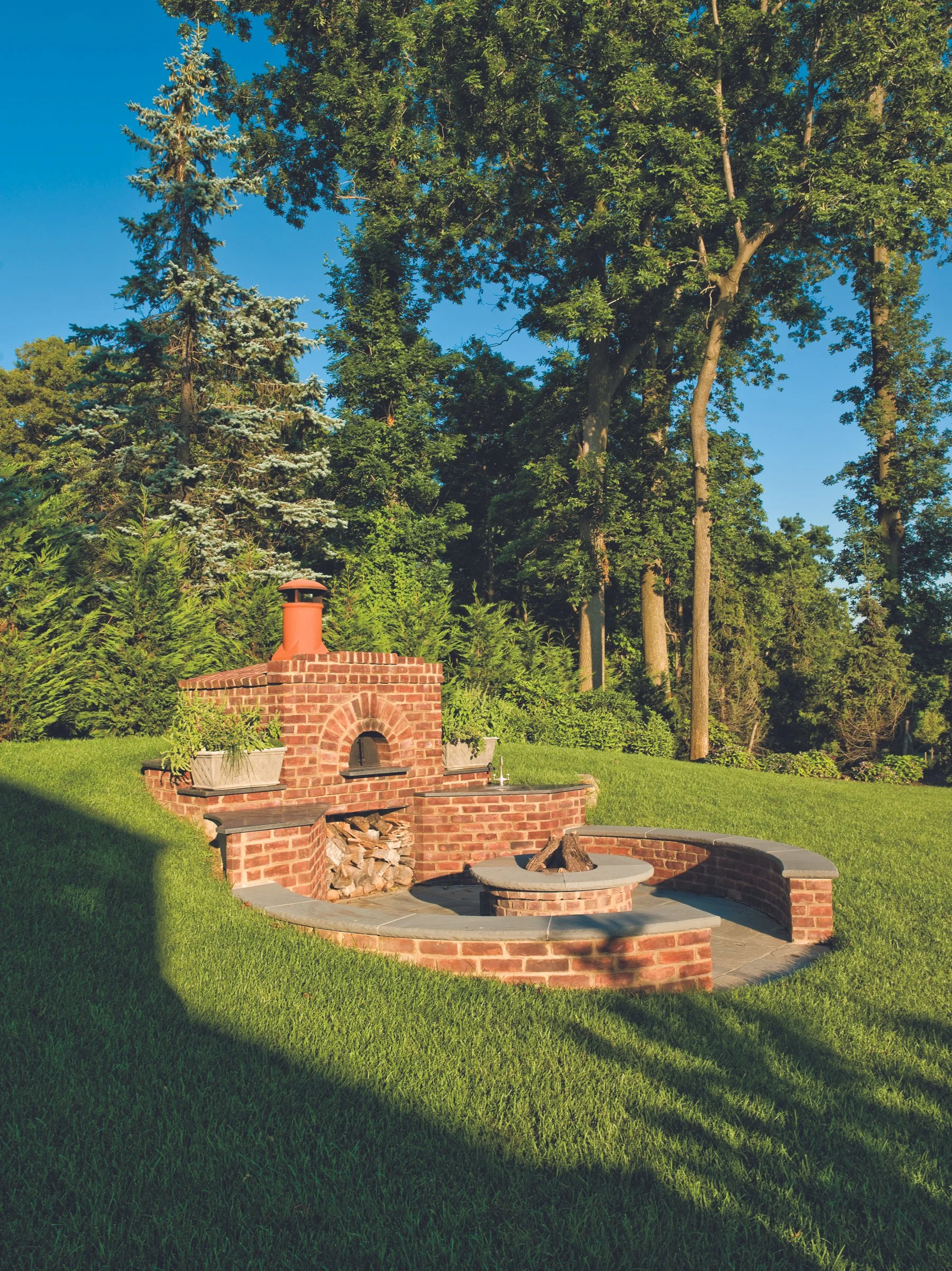 Outdoor brick fireplace with a chimney, surrounded by green grass and tall trees, under a clear blue sky.