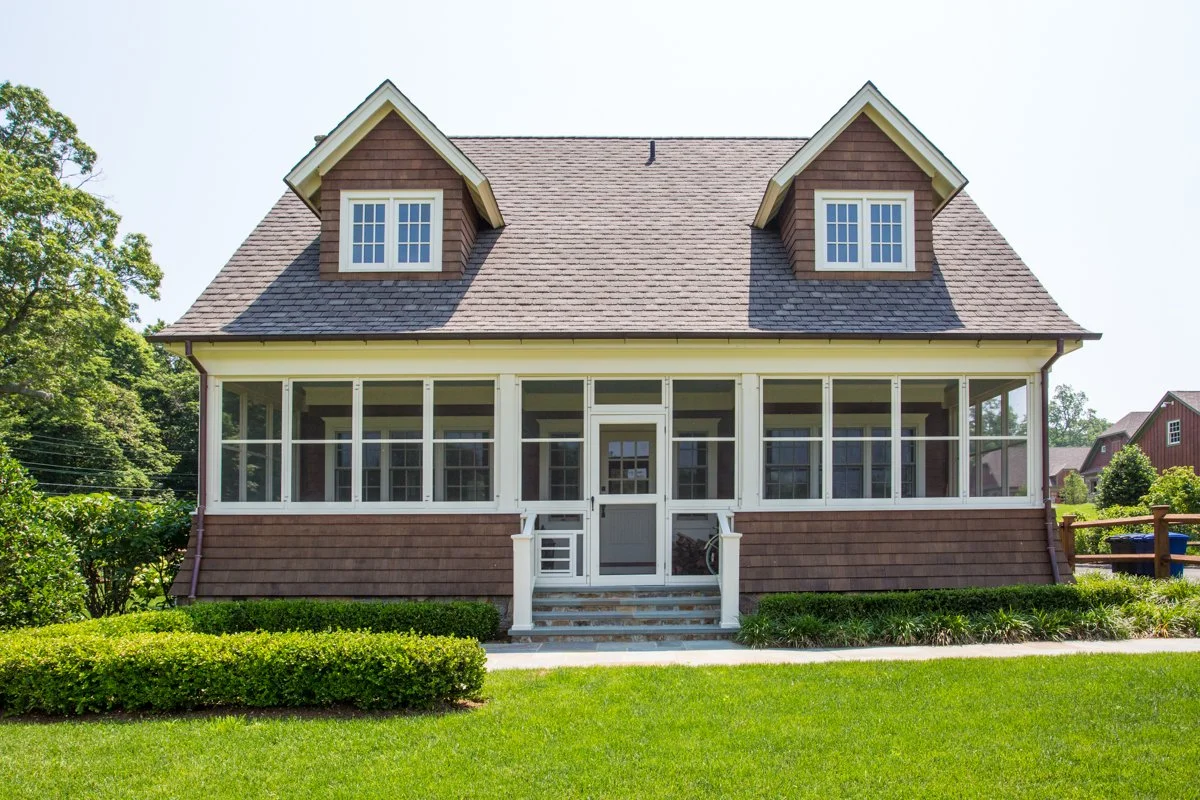 Two-story house with brown shingle siding, large screened porch, and dormer windows on a sunny day with green lawn and bushes in the front yard.