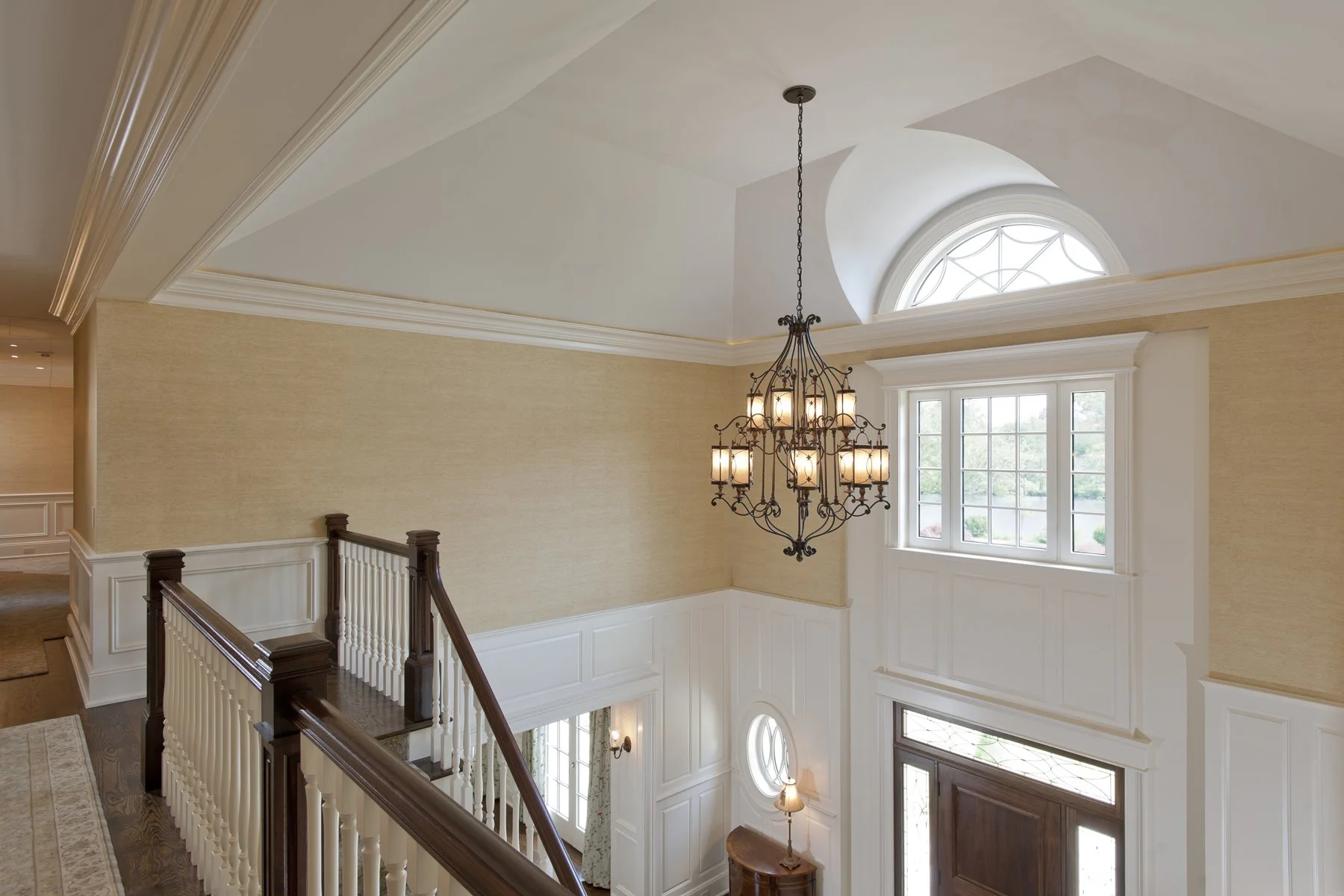 Interior of a house entryway featuring a high ceiling, a chandelier, a window with an arched top, and a wooden front door.