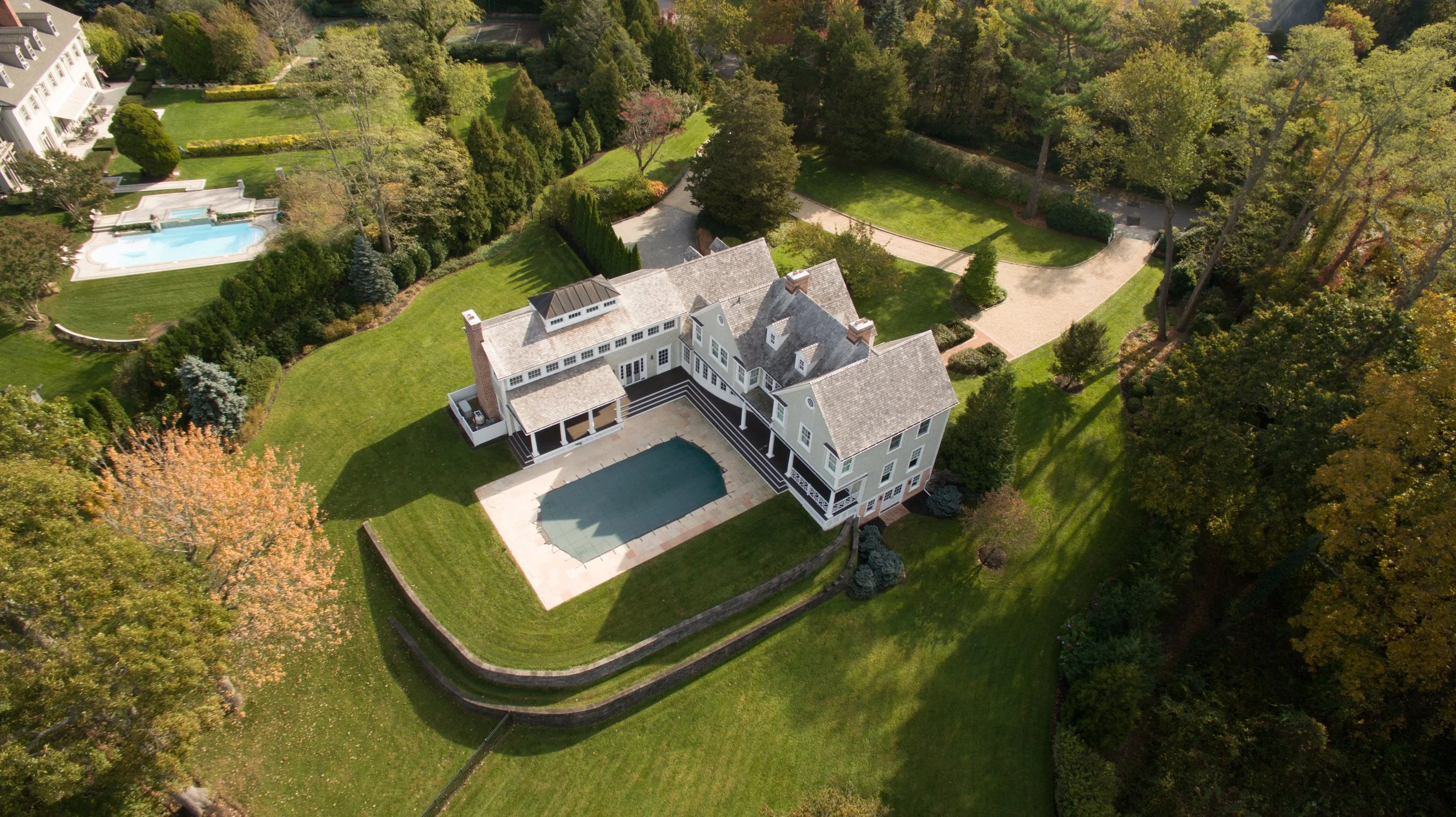 A large, multi-story house with a backyard swimming pool, surrounded by green lawns, trees, and pathways, with neighboring properties visible in the background.