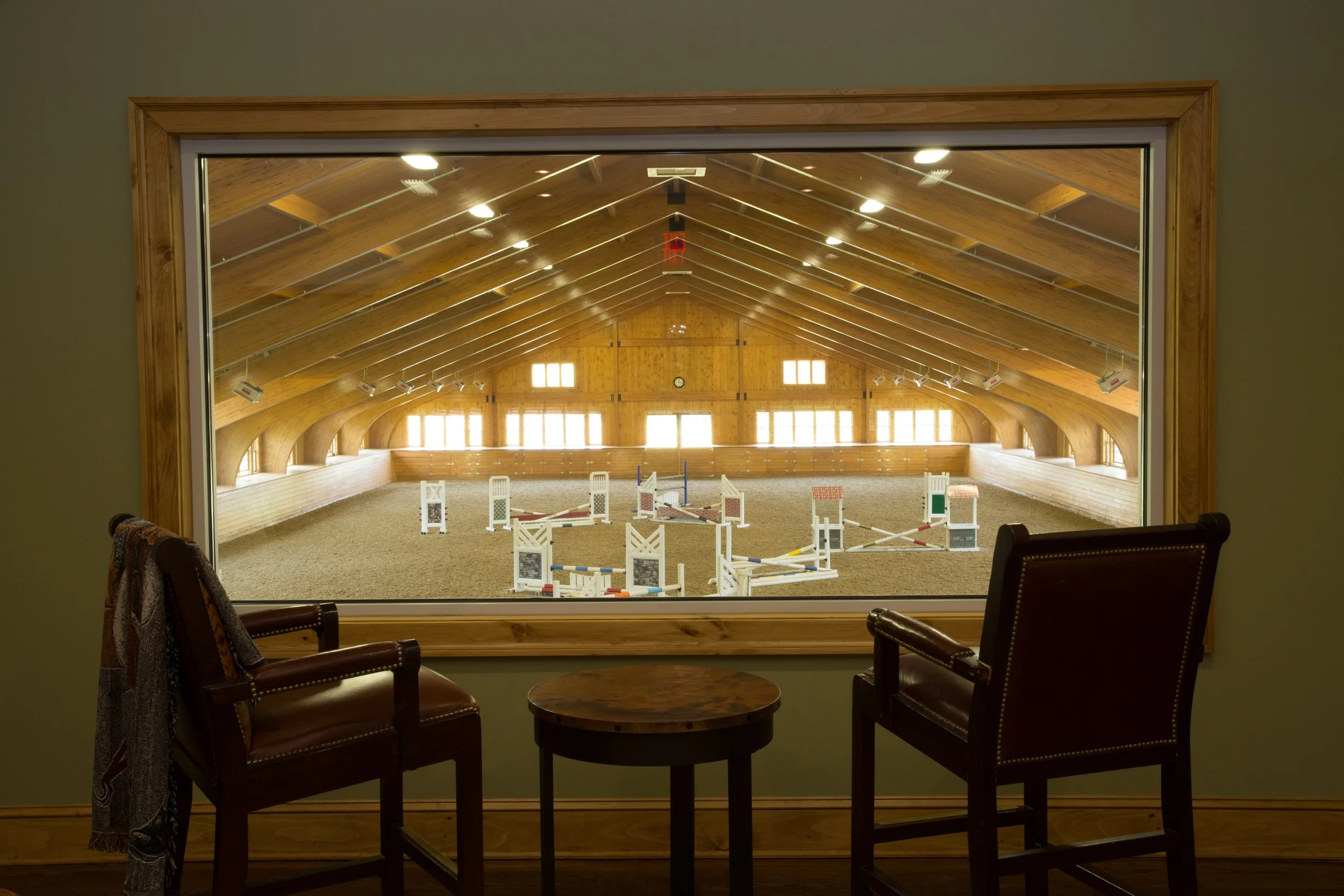 Indoor horse riding arena observed through a large window from a sitting area with two dark brown chairs and a small round table, wooden vaulted ceiling, and natural light.