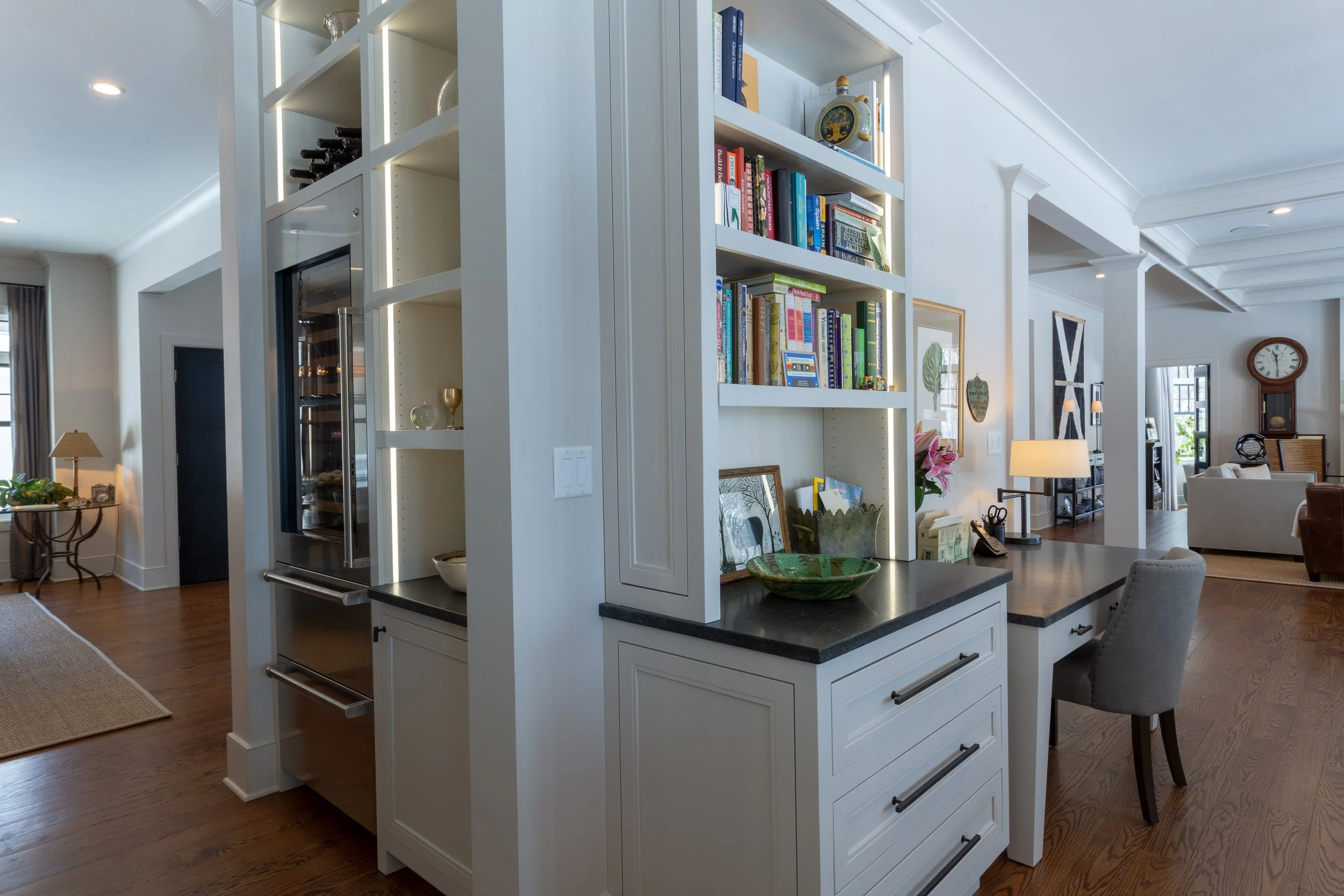 Open-plan living room with built-in white bookshelves, a kitchen counter, and seating area, with various books, decorations, and lighting fixtures.