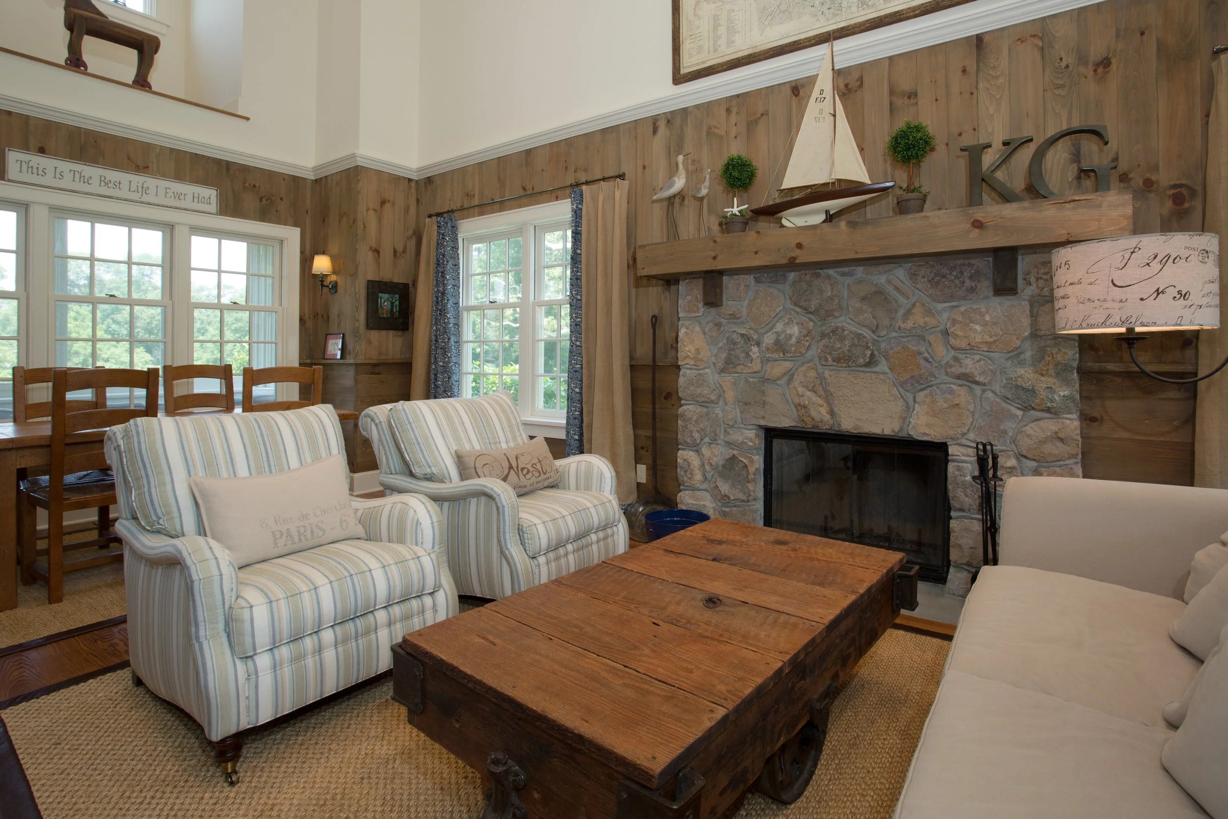 Cozy living room with a stone fireplace, wooden mantle, striped armchairs, a white sofa, rustic wooden coffee table, and decorative accents including potted plants, sailboat model, framed photos, and a sign reading "This Is The Best Life I Ever Had."