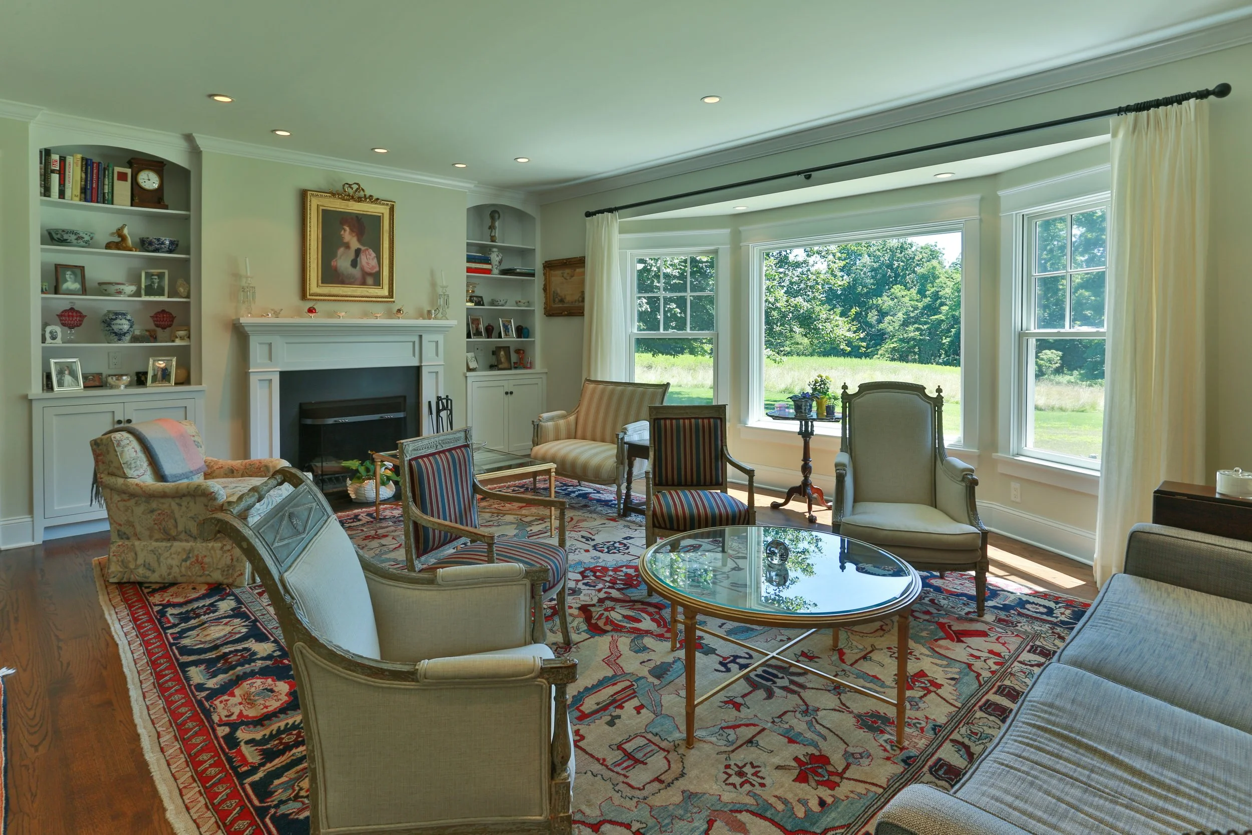 Living room with large windows, traditional furniture, a fireplace, and decorative items on shelves and mantel, illuminated by natural sunlight.