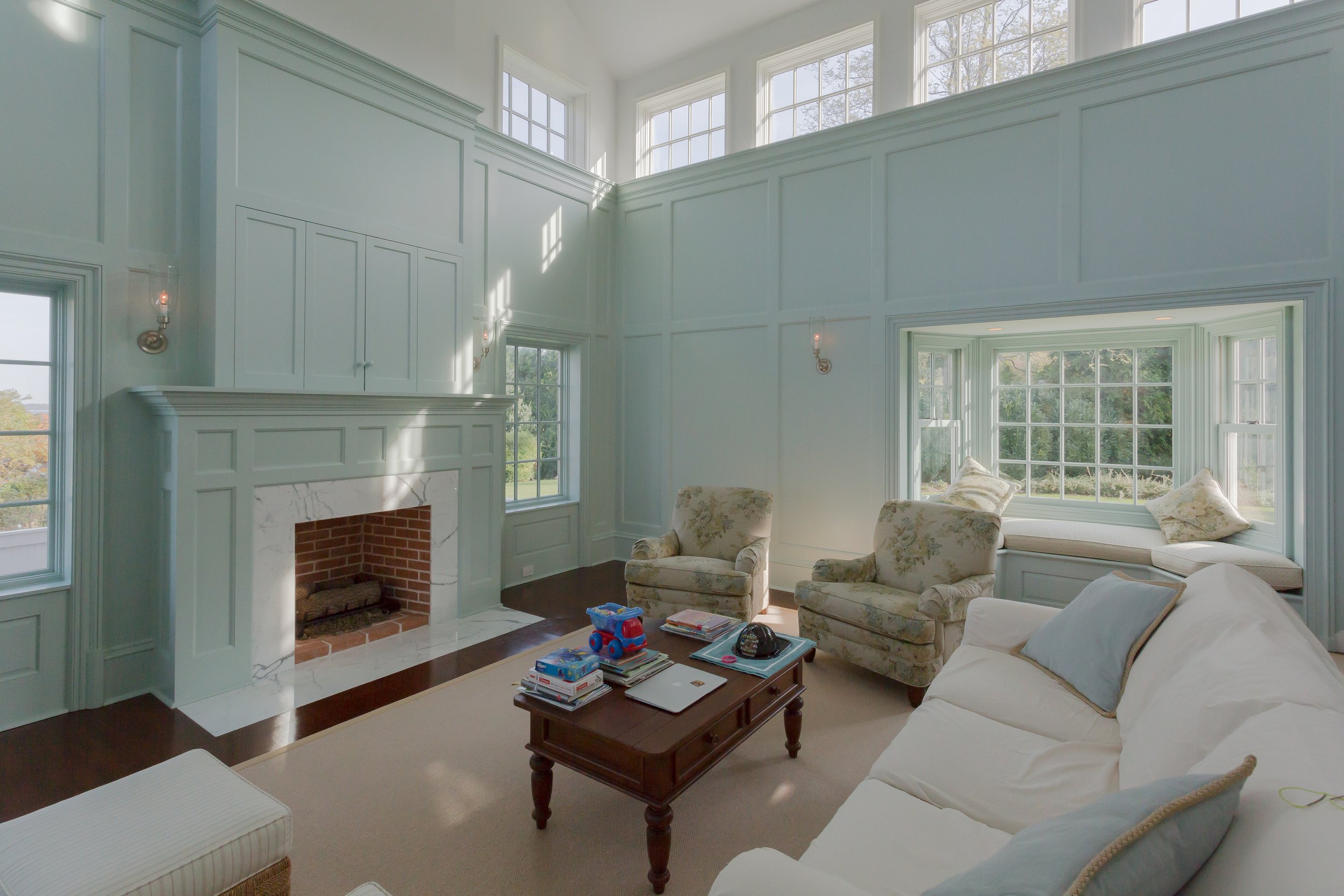 Living room with a high ceiling, large windows, a brick fireplace with white marble surround, beige armchairs, a white sofa with pillows, a wooden coffee table with children's toys and books, and a built-in window seat with cushions.