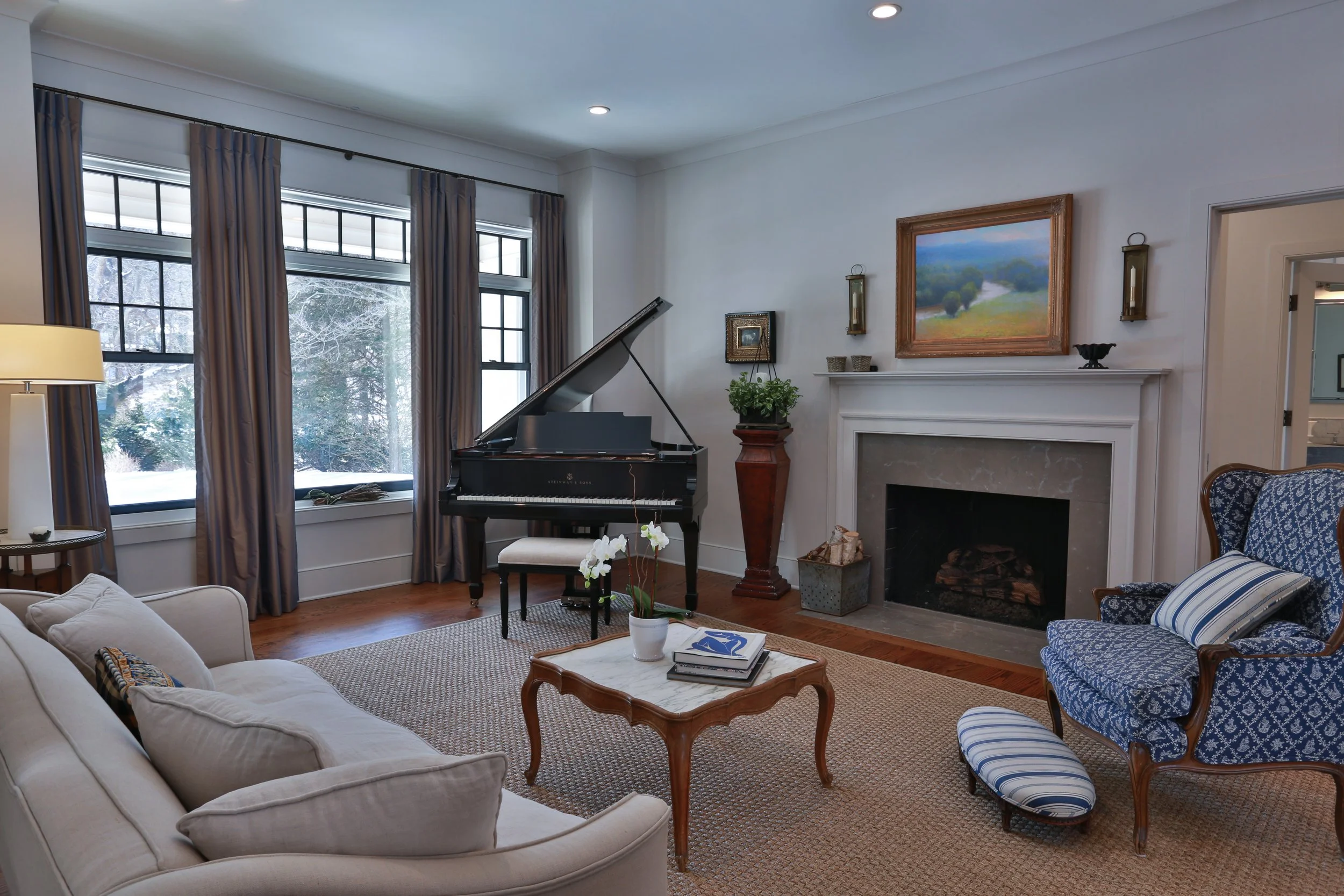 Living room with a grand piano, fireplace, blue patterned armchair, beige sofa, coffee table with magazines and a potted orchid, large windows with curtains, and a painting above the fireplace.