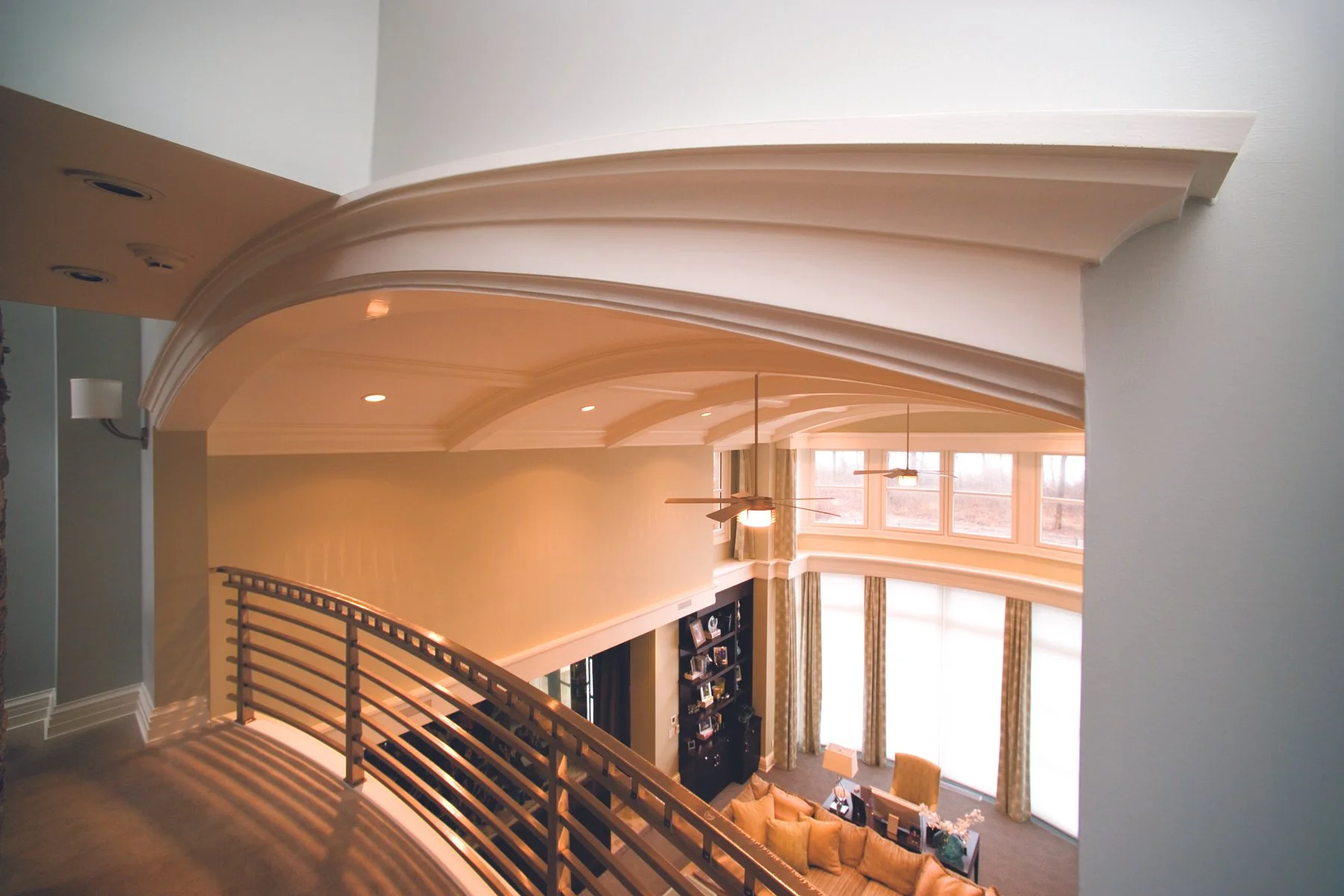 View from top of staircase overlooking a spacious living room with large bay windows, curtains, a ceiling fan, and furniture.
