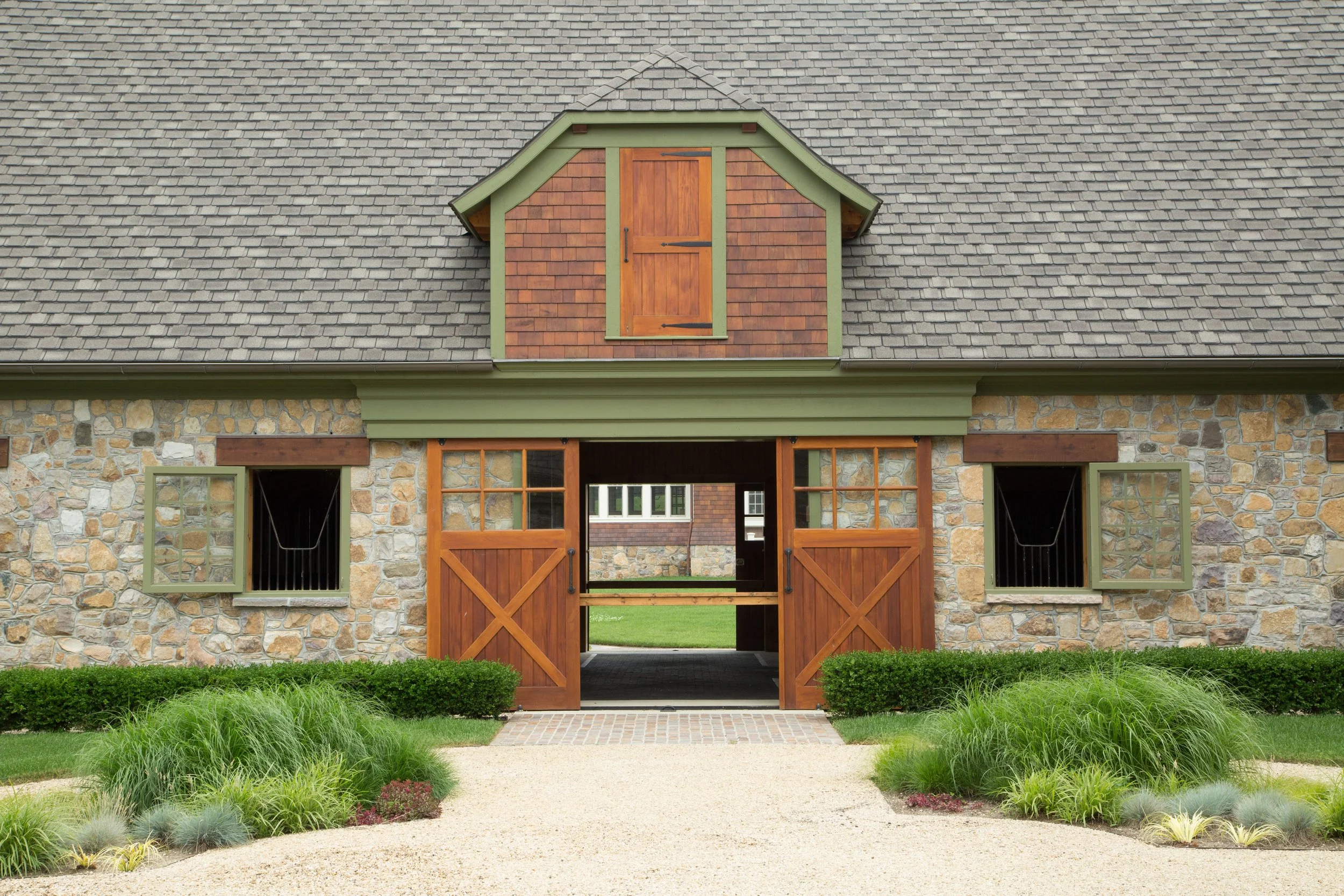 Front view of a stone and wooden barn with open doors, small windows, a shingled roof, and a landscaped pathway with green bushes.