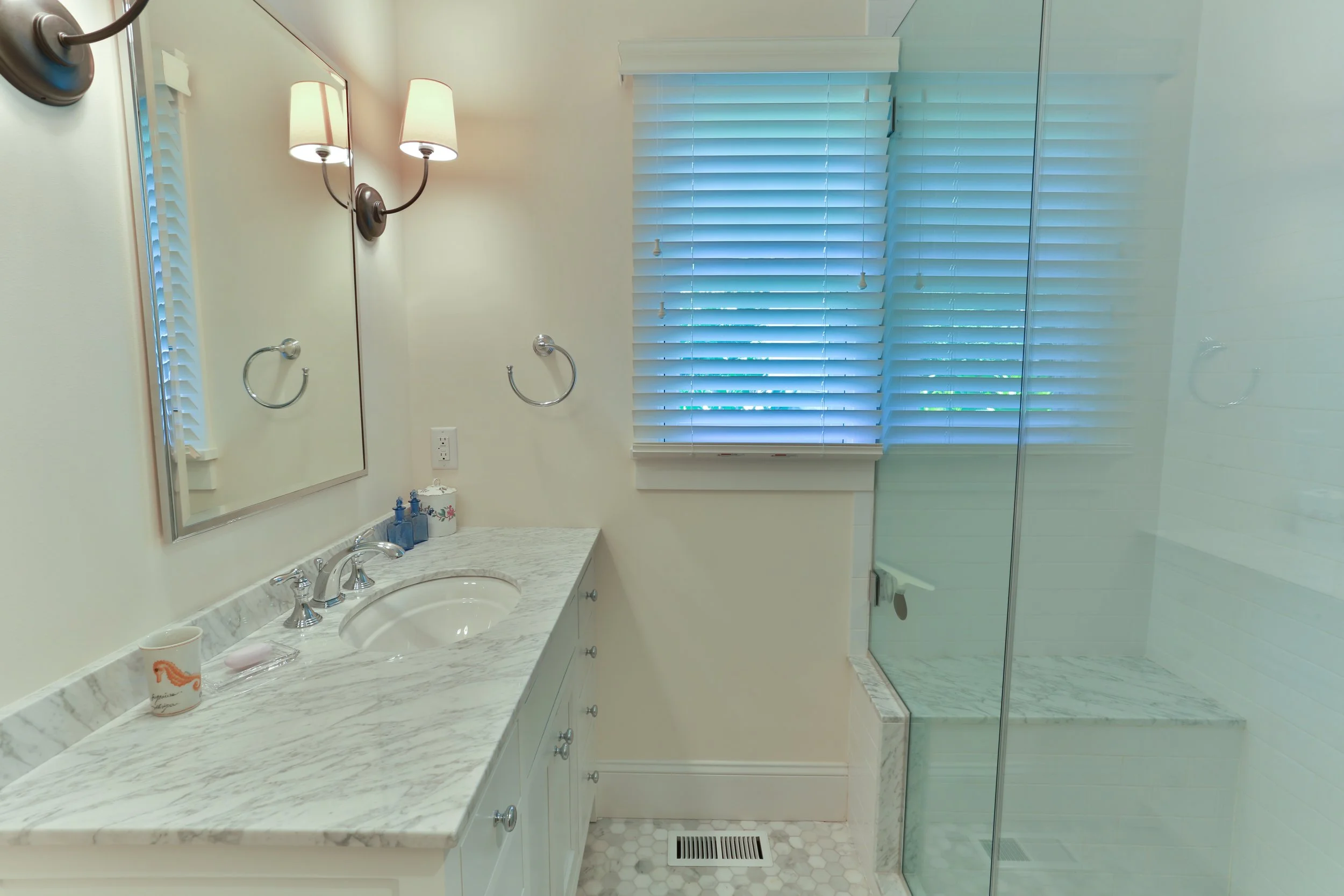 A bathroom with a marble countertop sink, a mirror, wall-mounted lights, and a walk-in shower with a glass door and white tiled walls. There are window blinds covering a window.