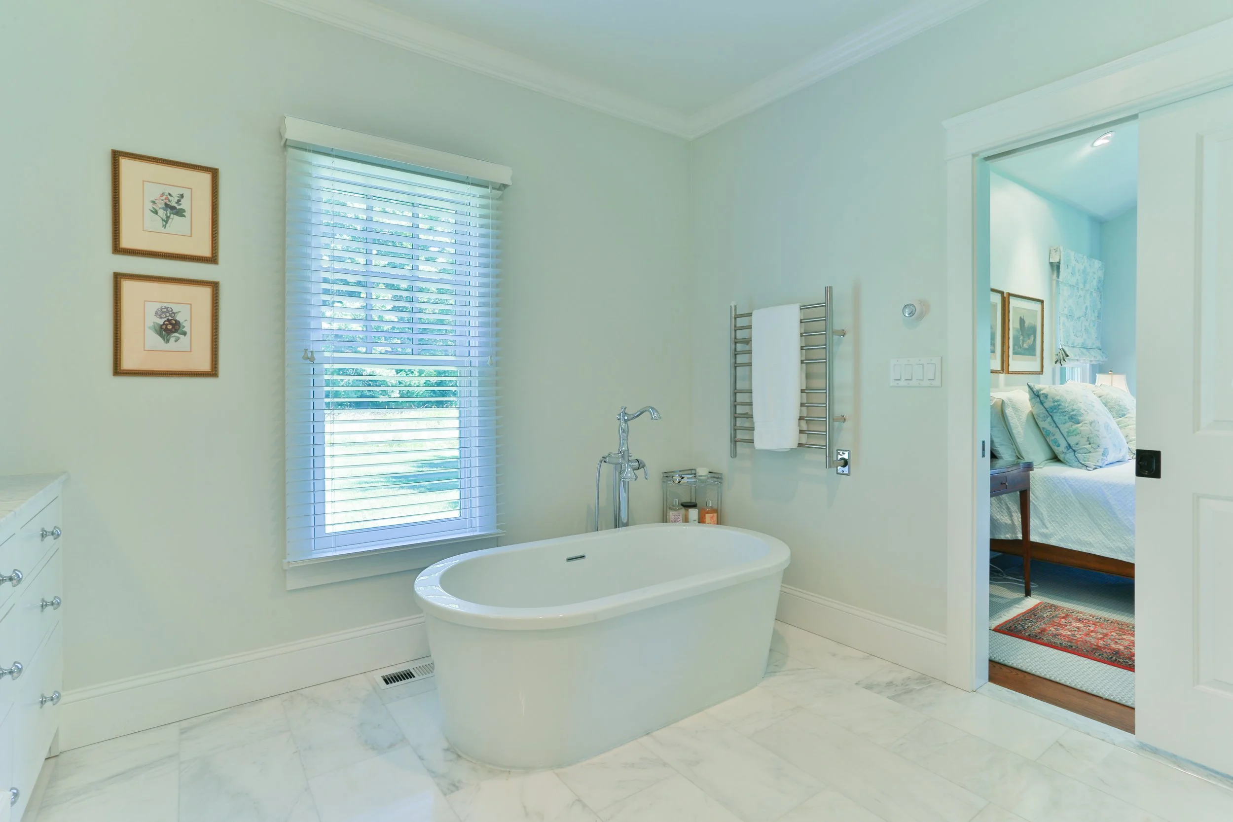 Bright bathroom with a white bathtub, window with blinds, framed botanical artwork, wall-mounted towel rack, and door leading to bedroom with floral bedding.