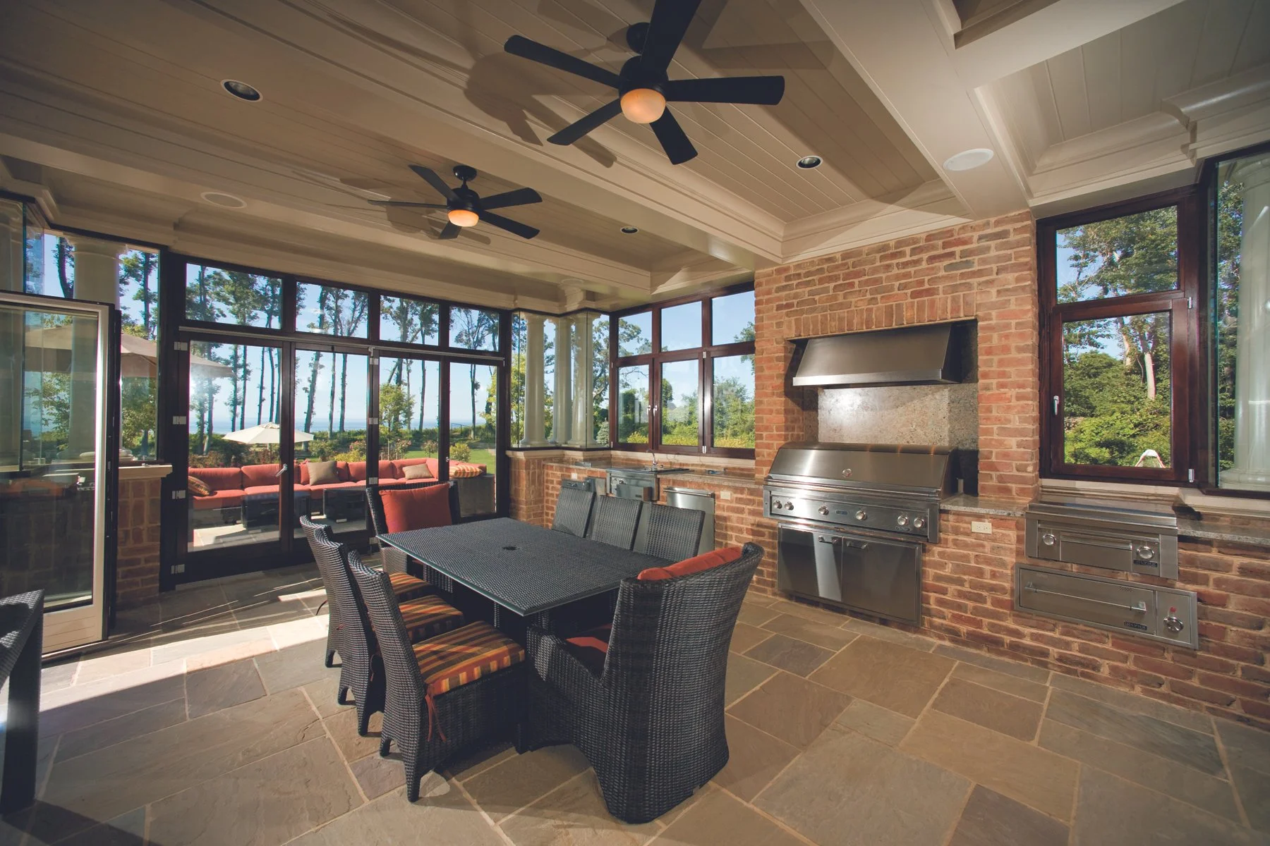 Indoor patio with brick walls, large windows, ceiling fans, a dining table with six chairs, and a built-in stainless steel grill.
