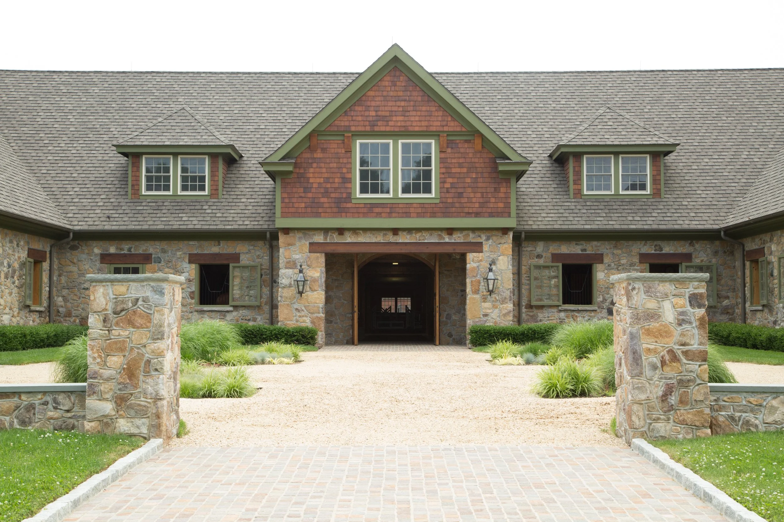 A large house with a stone exterior, green trim, and a gray shingle roof. The house features dormer windows and a central arched entrance with lanterns on each side.