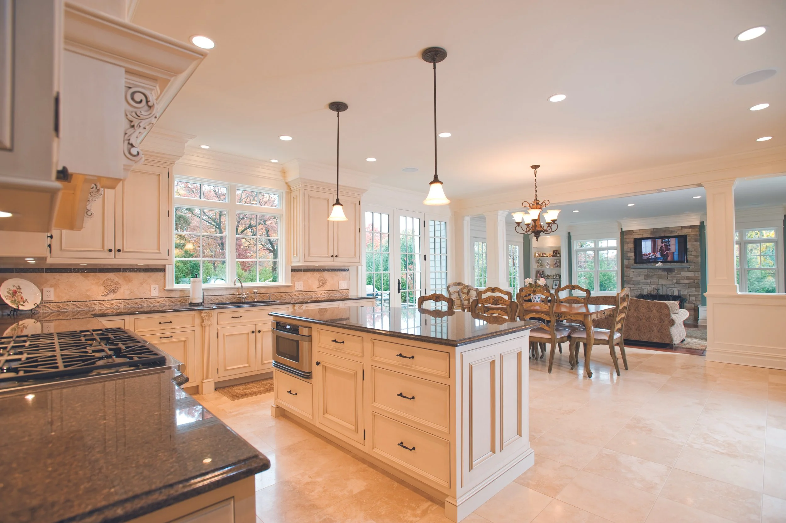 Spacious open-concept kitchen and dining area with cream-colored cabinetry, granite countertops, large central island, stainless steel appliances, pendant lights, and a view into the living room with a stone fireplace and flat-screen TV.