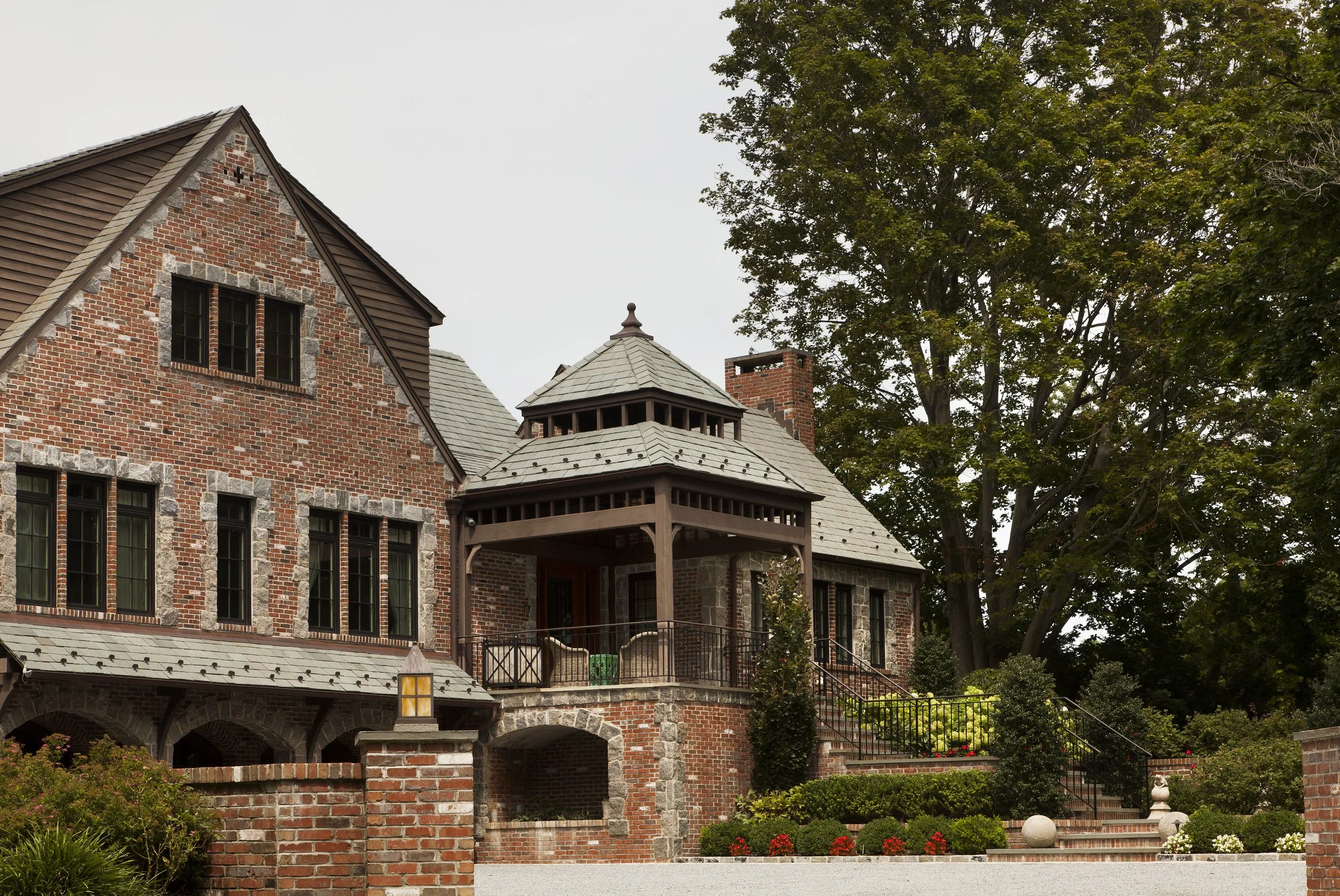 A large brick house with a steep roof, chimney, and decorative stone accents, surrounded by landscaped gardens and trees.