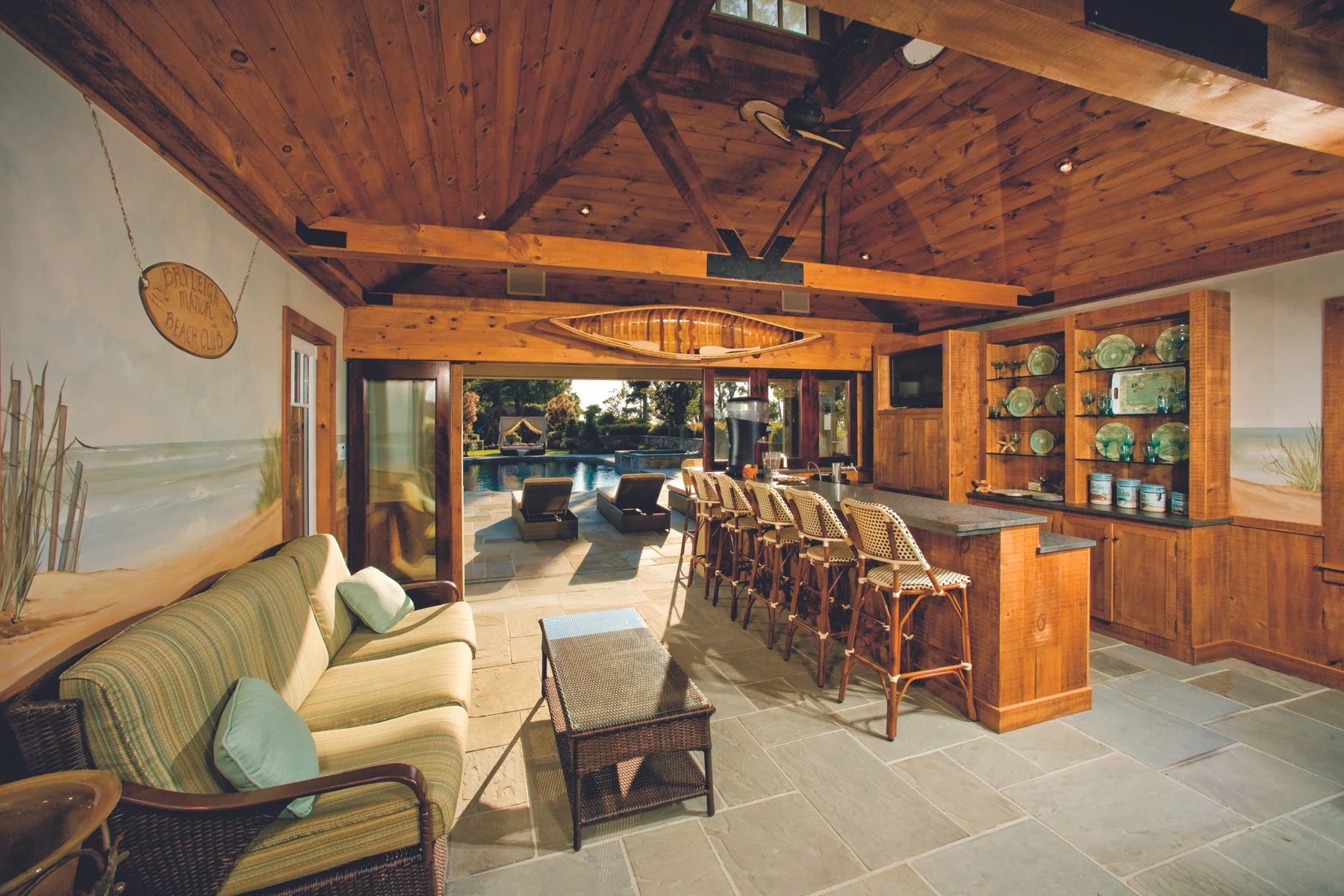 Indoor living space with a bar counter, wicker chairs, a sofa with green cushions, and a view of an outdoor pool area with lounge chairs.