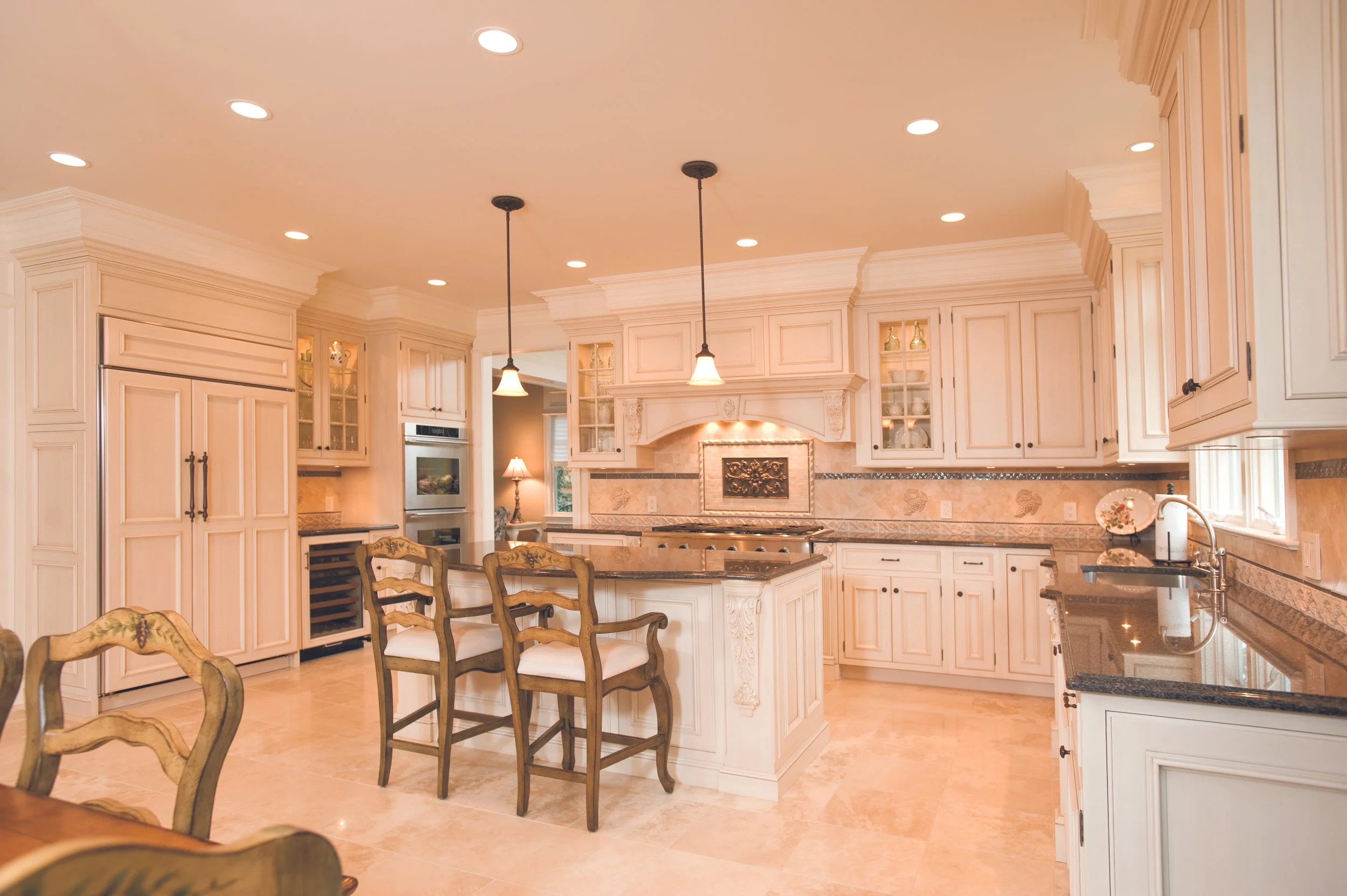 Bright and spacious kitchen with cream-colored cabinets, black granite countertops, and beige tile flooring. There is a central island with chairs, pendant lighting, and built-in appliances. The kitchen has decorative molding and a picture on the wal